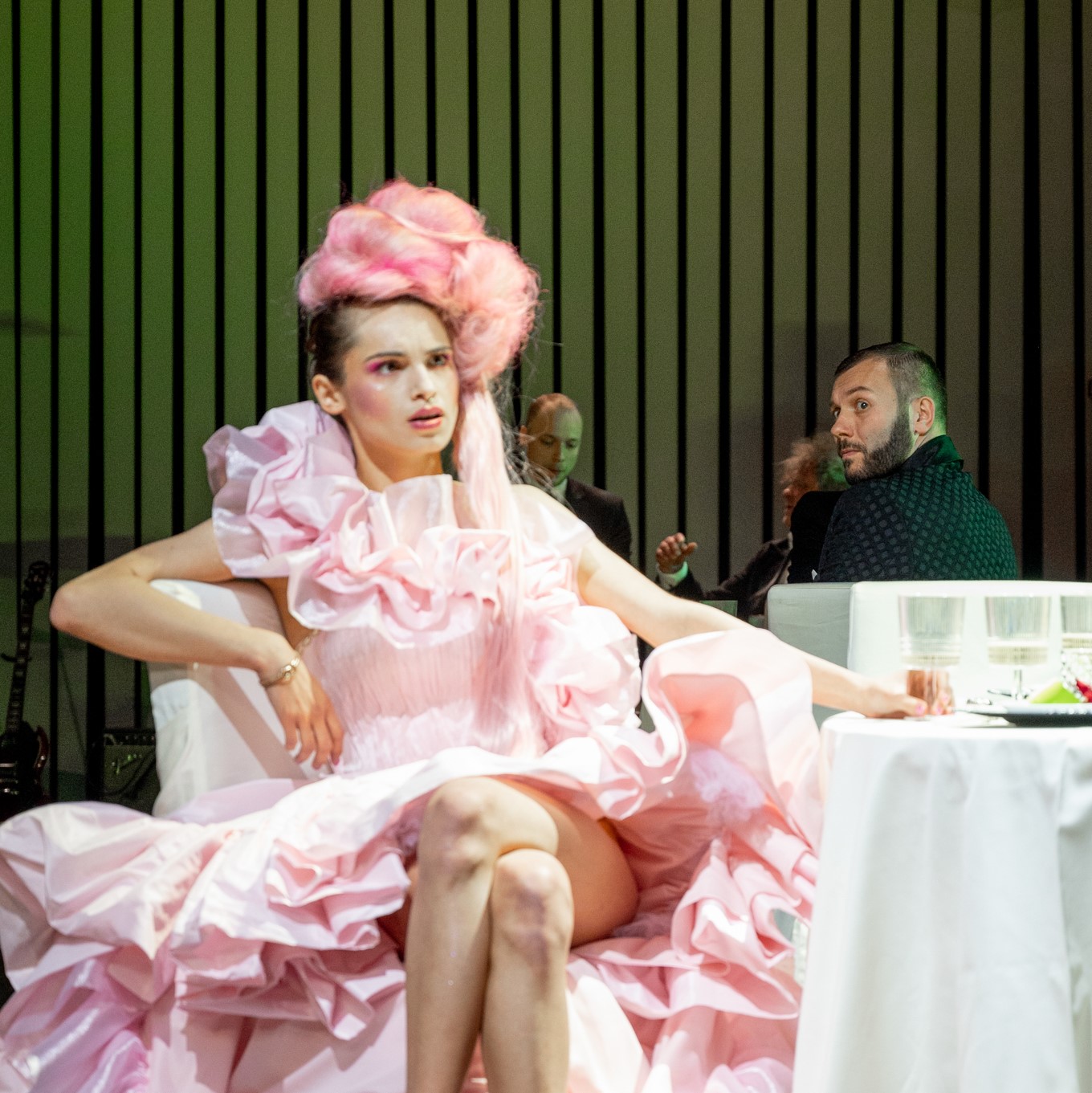 Aktorka w strojnej różowej sukni i różowej peruce doczepionej na szczycie głowy siedzi przy stole z nogą założoną na nogę.
