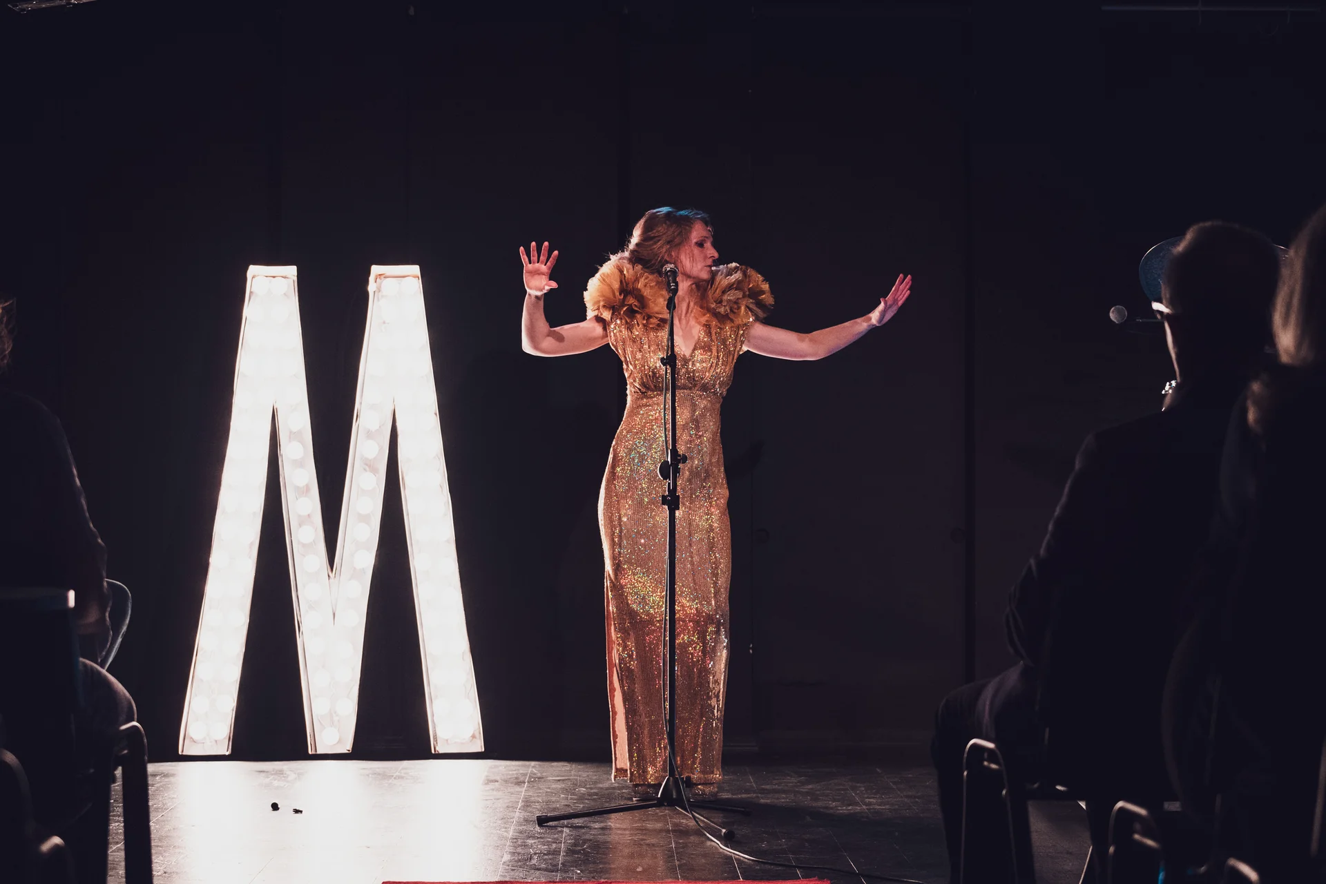 Maria w złotej sukni z uniesionymi rękami stoi przed mikrofonem. Za nią świecącą gigantyczna litera "M".