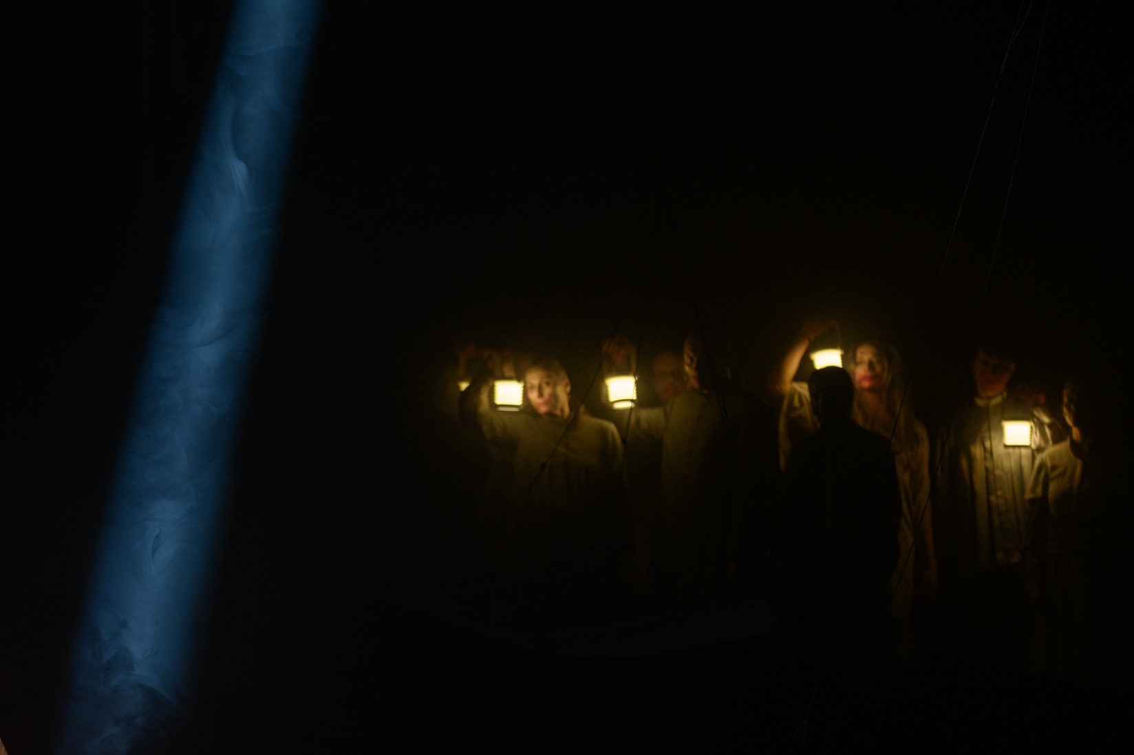 Z ciemności wyłaniają się osoby o trupich twarzach, trzymając w rękach lampy naftowe.
