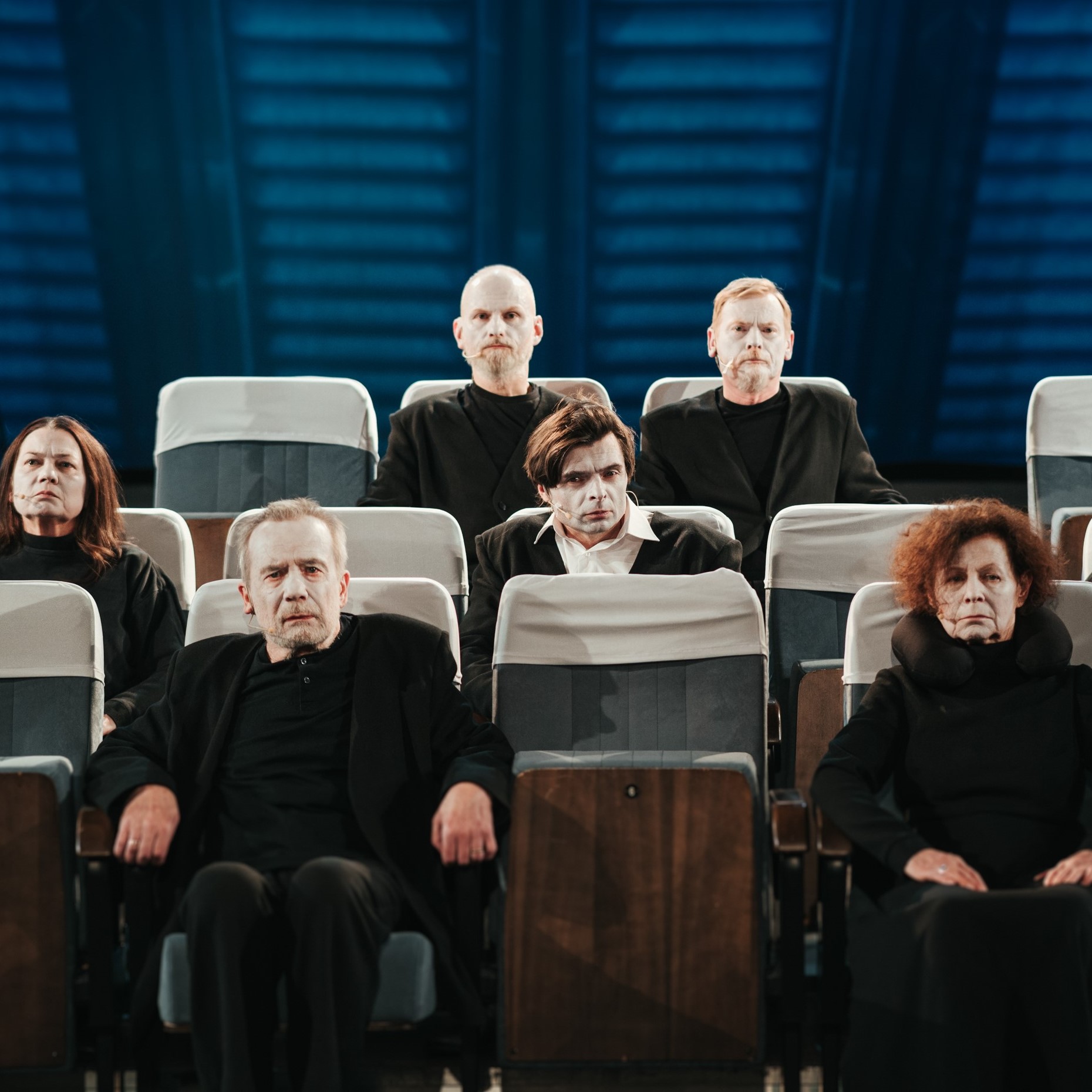 Niebieskie tło, trzy rzędy foteli, na których siedzą aktorzy.