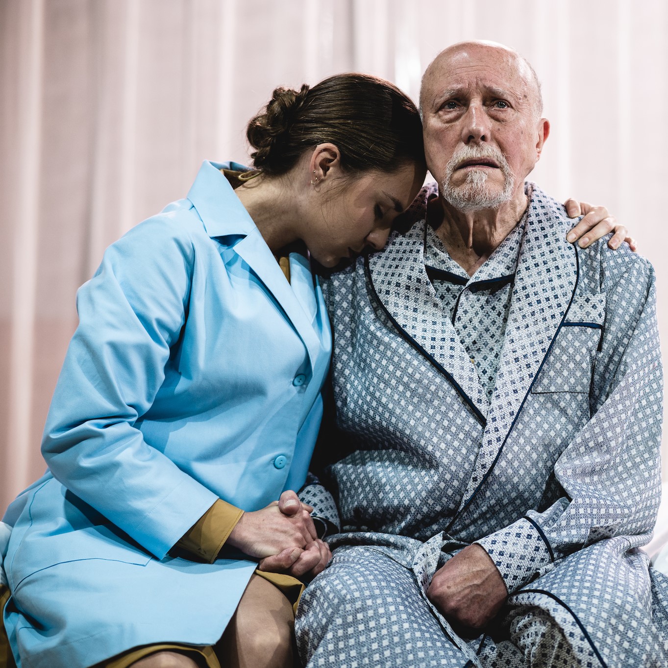 Starszy mężczyzna siedzi i smutno wpatruje się w przestrzeń. Obejmuje go młoda kobieta w stroju pielęgniarskim.
