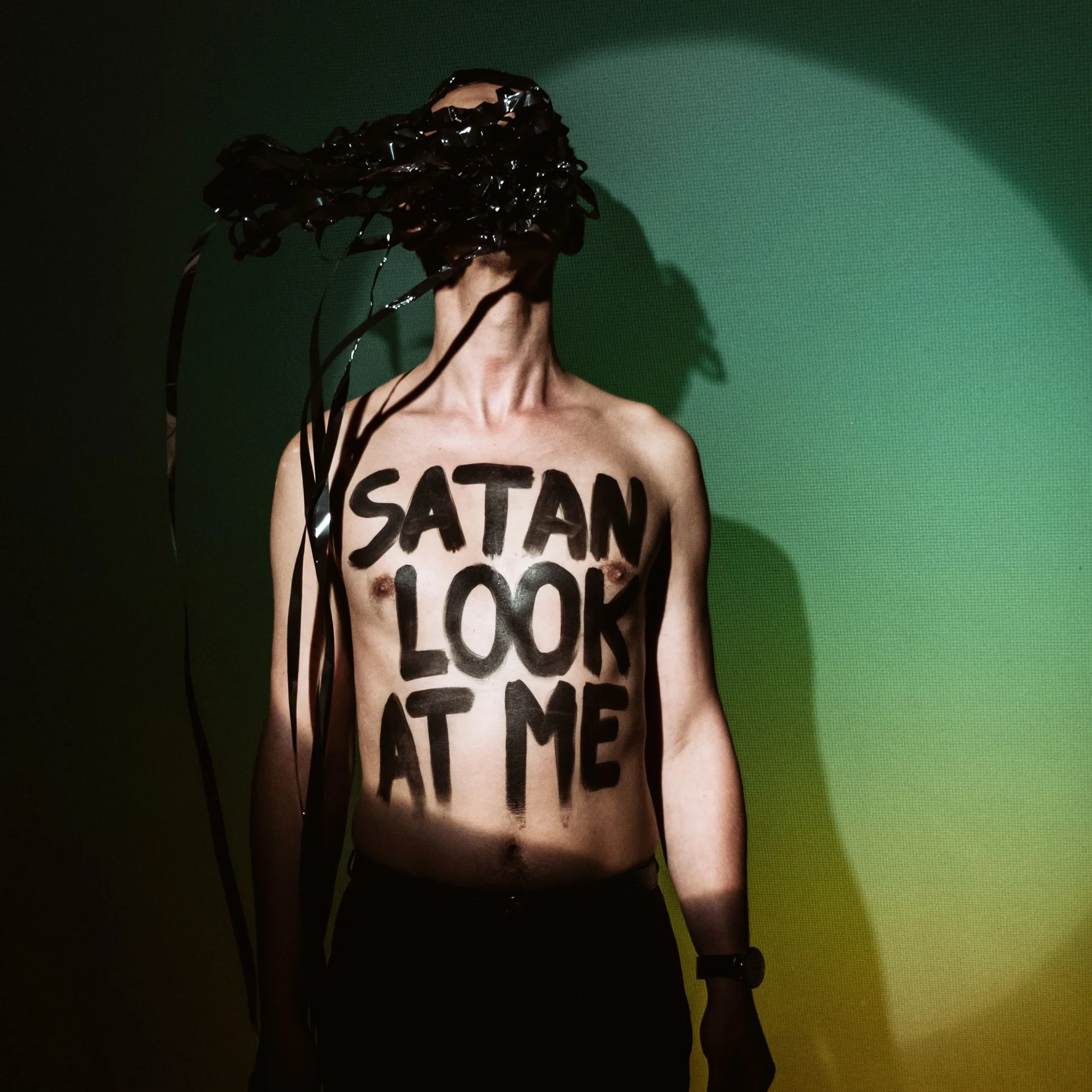 Mężczyzna bez koszulki trzyma w zębach kłębek czarnej taśmy magnetofonowej. Na klatce piersiowej ma napis: "Satan look at me".
