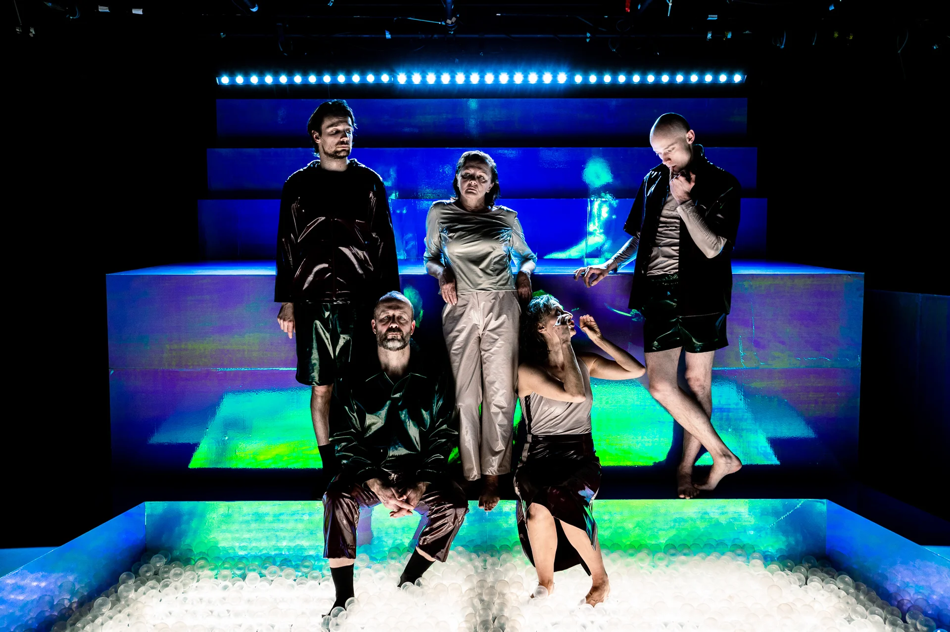 Grupa aktorów w skórzanych kostiumach siedzi na podświetlonych elementach scenografii.