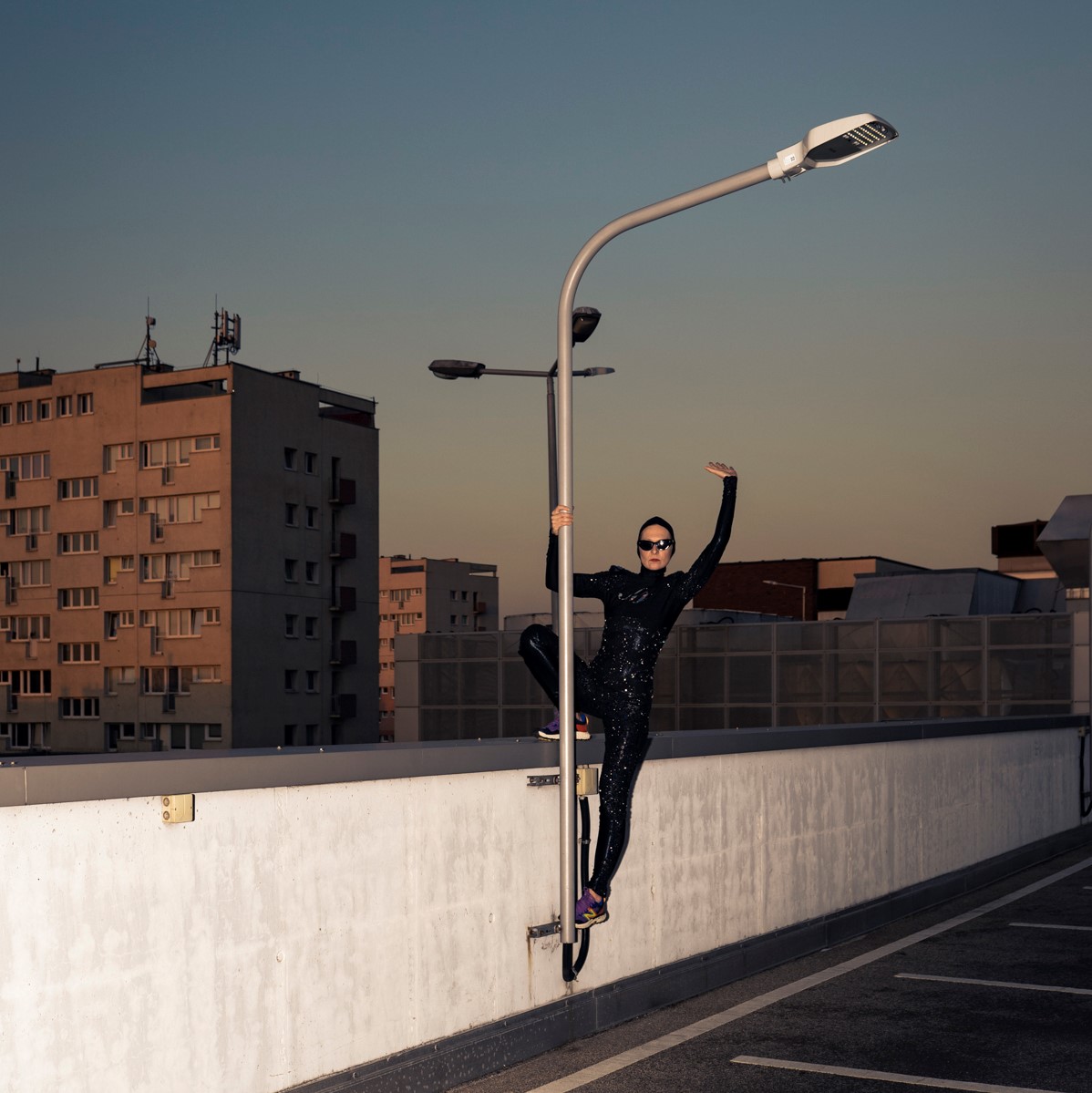 Kobieta w czarnym obcisłym kostiumie wspina się na latarnię miejską. Jedną rękę unosi do góry. W tle widoczne wieżowce.