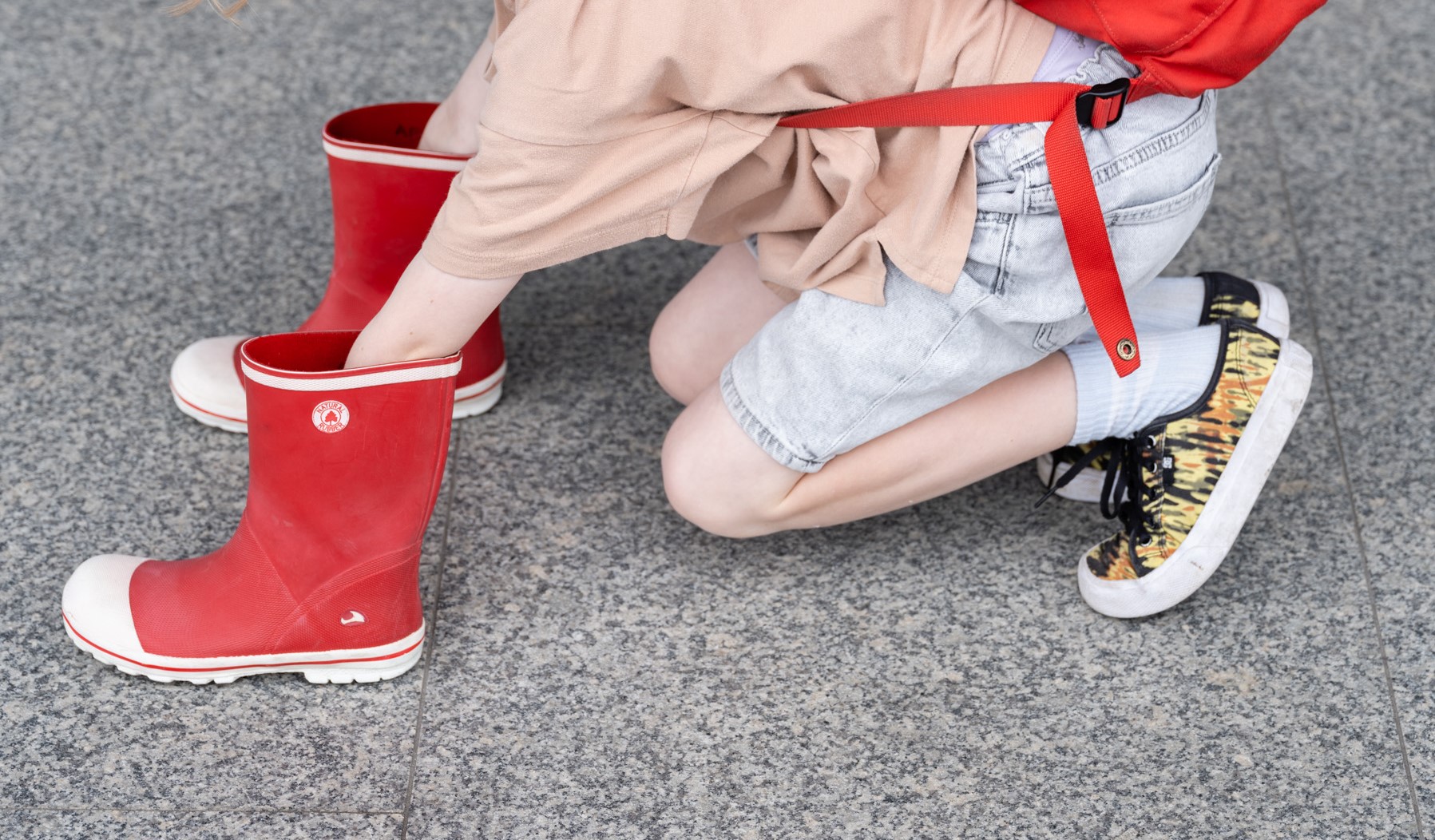 Dziewczynka z czerwonym plecakiem kuca i wsadza dłonie w czerwone kalosze.