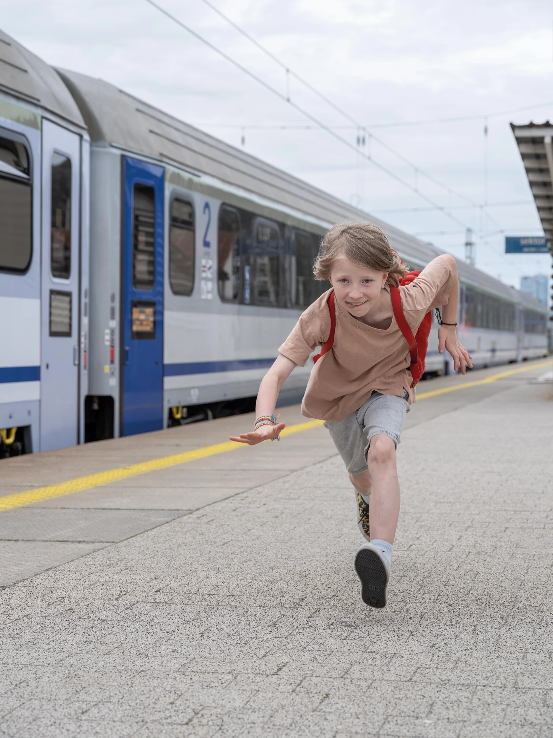 Dziewczynka z czerwonym plecakiem biegnie po peronie kolejowym. W tle stojący pociąg.