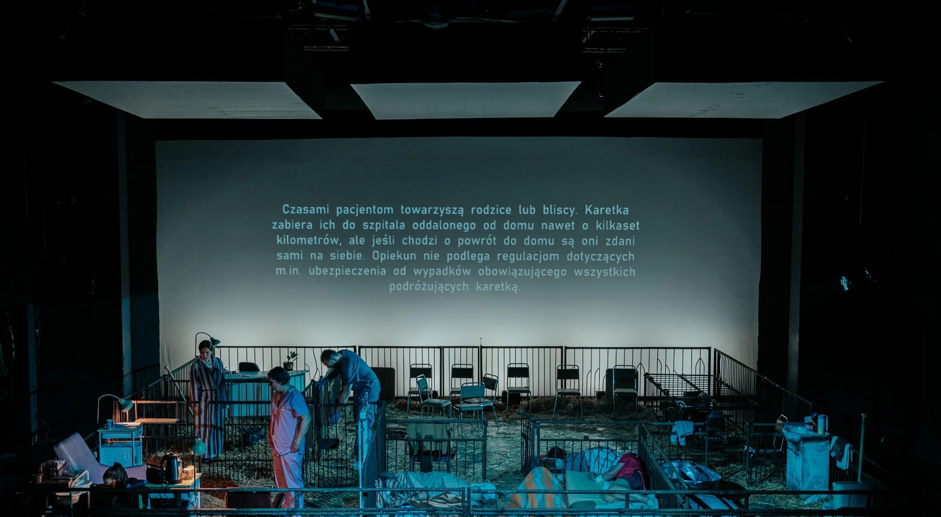 Zdjęcie z oddalenia, scenografia spektaklu "Spartakus. Miłość w czasach zarazy". Widoczne metalowe wygrodzenia, pomiędzy nimi szpitalne meble, ludzie w kitlach i pidżamach, na ścianie wyświetlany napis.