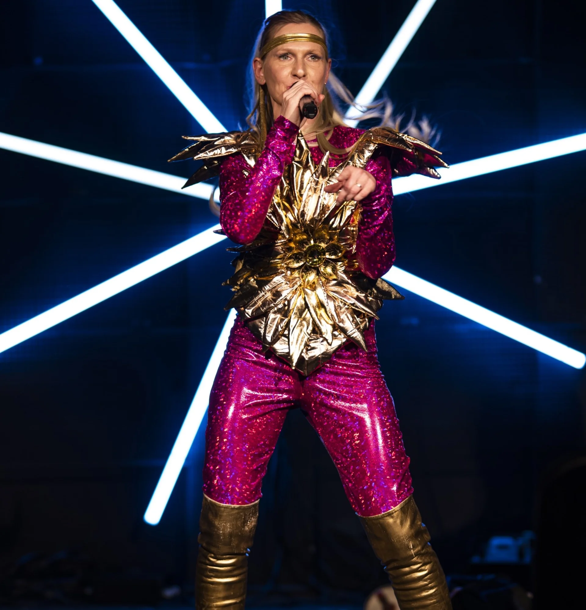 Kobieta w różowo-złotym scenicznym kostiumie śpiewa do mikrofonu. Za nią widoczny neon.