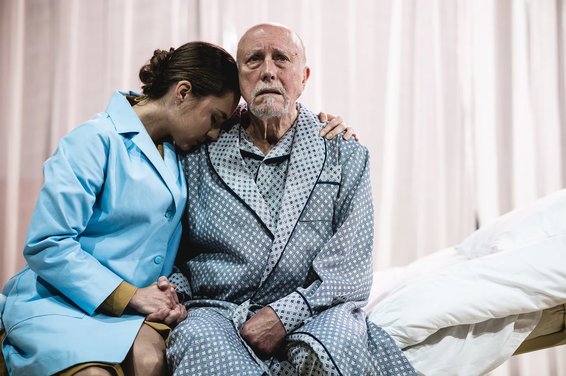 Siedzący na łóżku starszy aktor w piżamie z przerażoną miną. Obok niego siedzi młodsza aktorka w błękitnej marynarce, trzyma go za dłoń i wtula w niego głowę.
