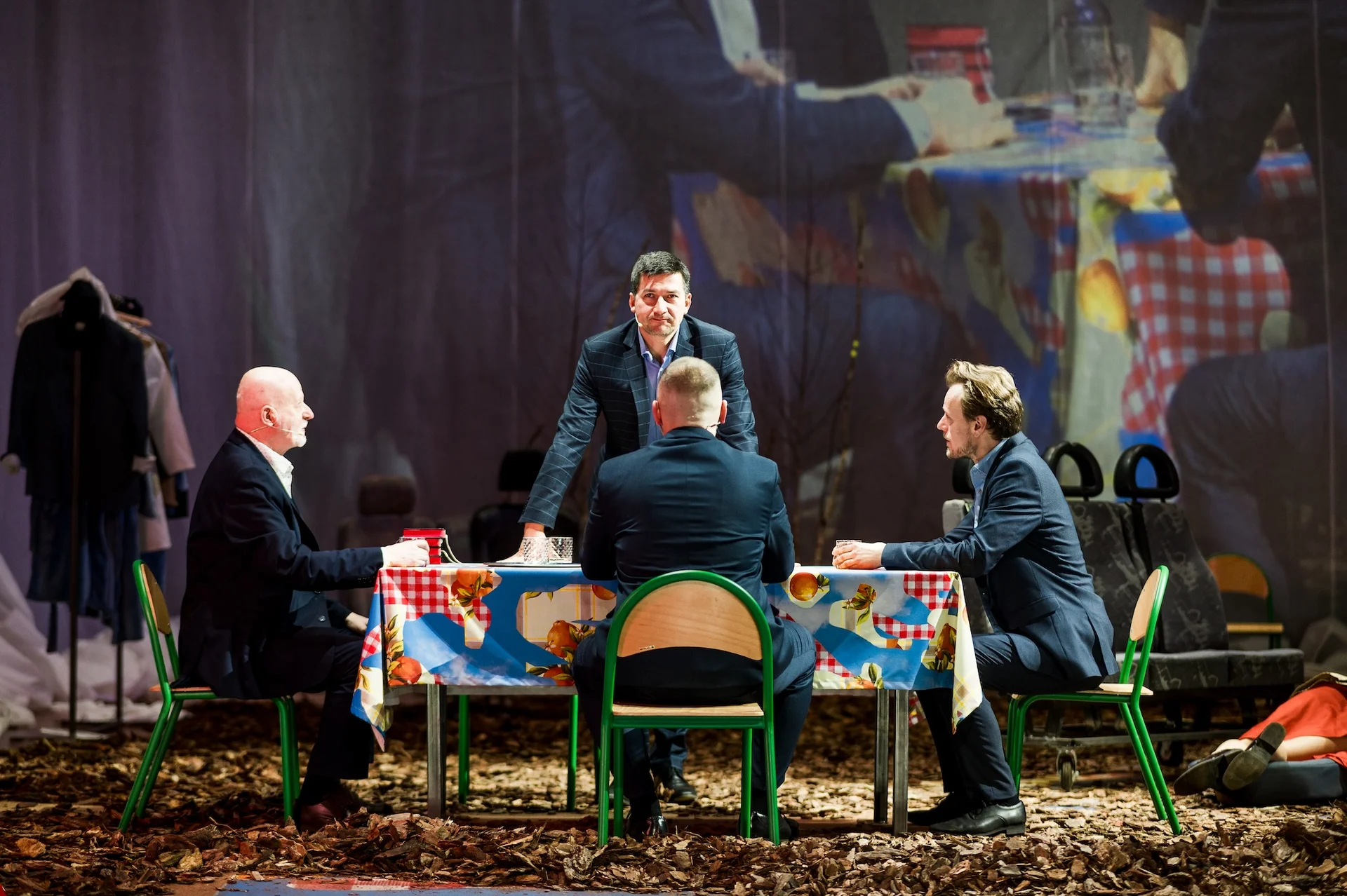 Czterech aktorów w garniturach siedzących przy stole. Jeden z nich wstał, oparł ręce na stole i patrzy na publiczność.