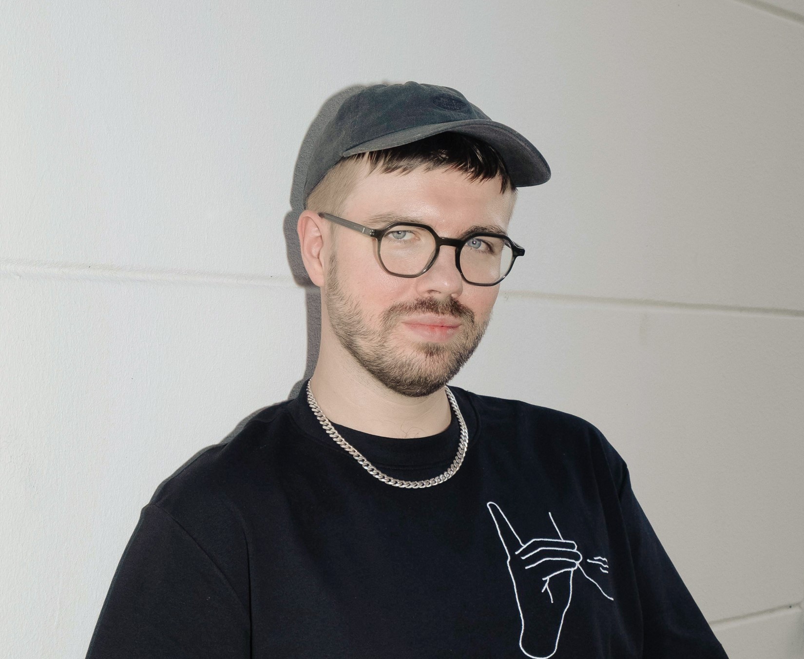Portret Daniela Kotowskiego. Młody mężczyzna w okularach, w czarnym t-shircie i czapce z daszkiem na tle białej ściany. 