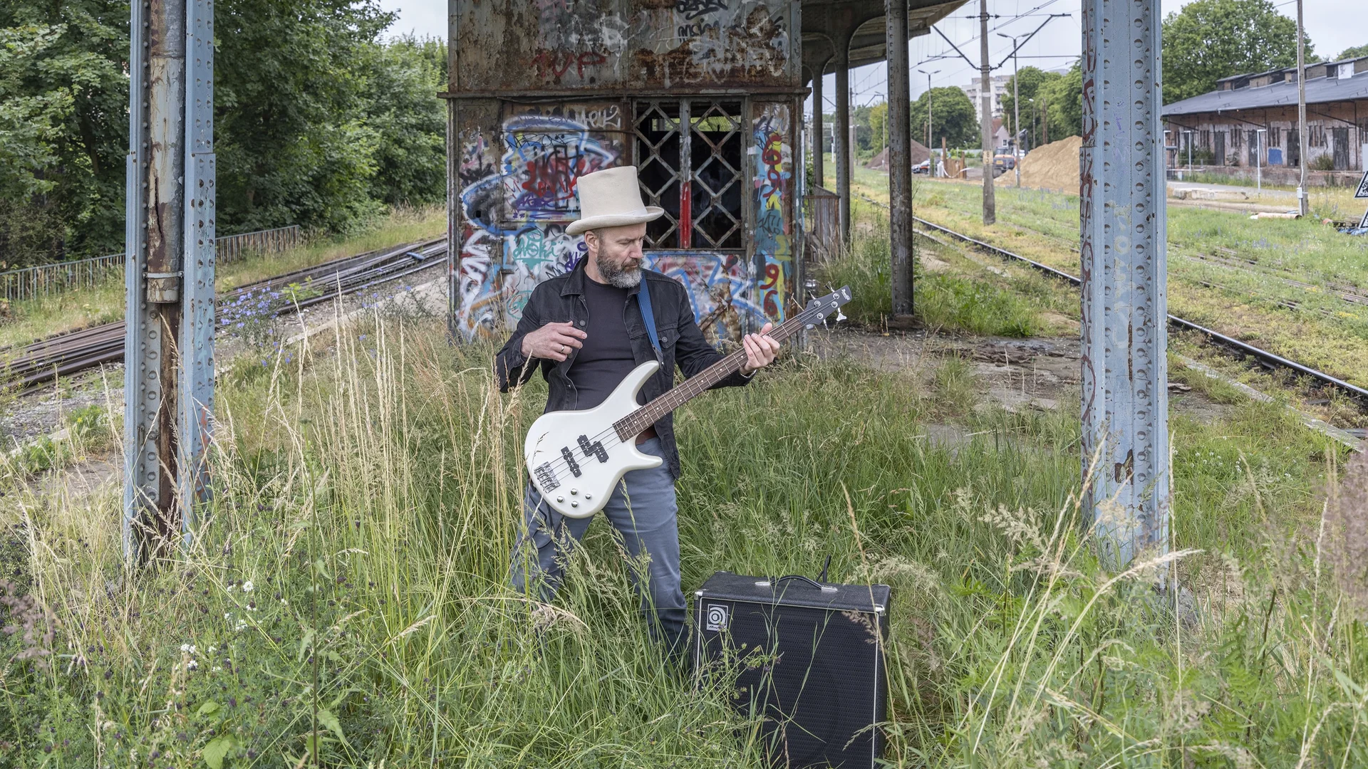 Mężczyzna w cylindrze gra na gitarze elektrycznej stojąc na starym dworcu.