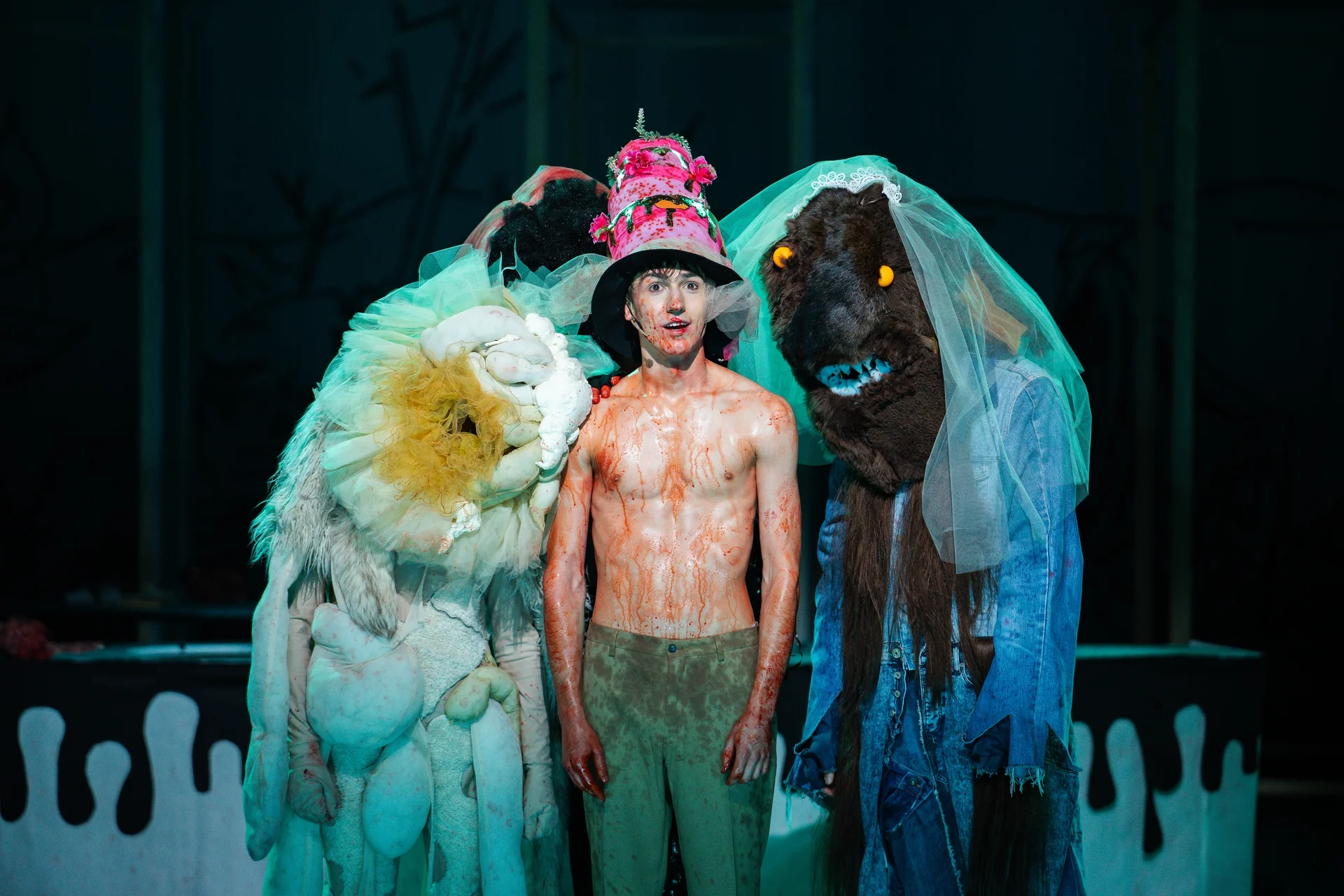 Młody mężczyzna z gołym torsem, oblany krwią. Na głowie ma kolorową, wysoką czapkę. Po jego bokach stoją włochate potwory.