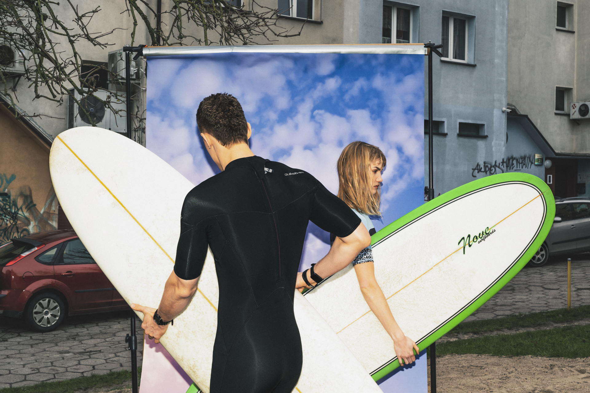 Dwoje serferów w piankach trzyma deski surfingowe. Serferzy (kobieta i mężczyzna) kierują swoje deski w przeciwnych kierunkach. Kobieta widoczna jest bokiem, mężczyzna tyłem. Oboje stoją na tle ekranu w chmury. W tle widoczne bloki.