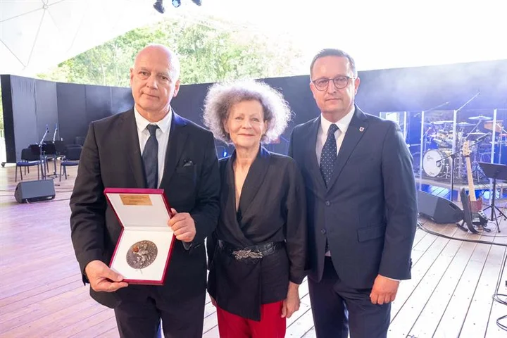 Zdjęcie z wręczenia nagrody Annie Januszewskiej. Scena teatru letniego w Szczecinie. W środku aktorka Anna Januszewska, w czarnym żakiecie i czerwonych spodniach. Po obu jej stronach mężczyźni w garniturach - wicemarszałek Bogdan Jaroszewicz i Roman Ciepliński, redaktor naczelny „Kuriera Szczecińskiego”. Jeden z mężczyzn trzyma pudełko z odznaczeniem. 