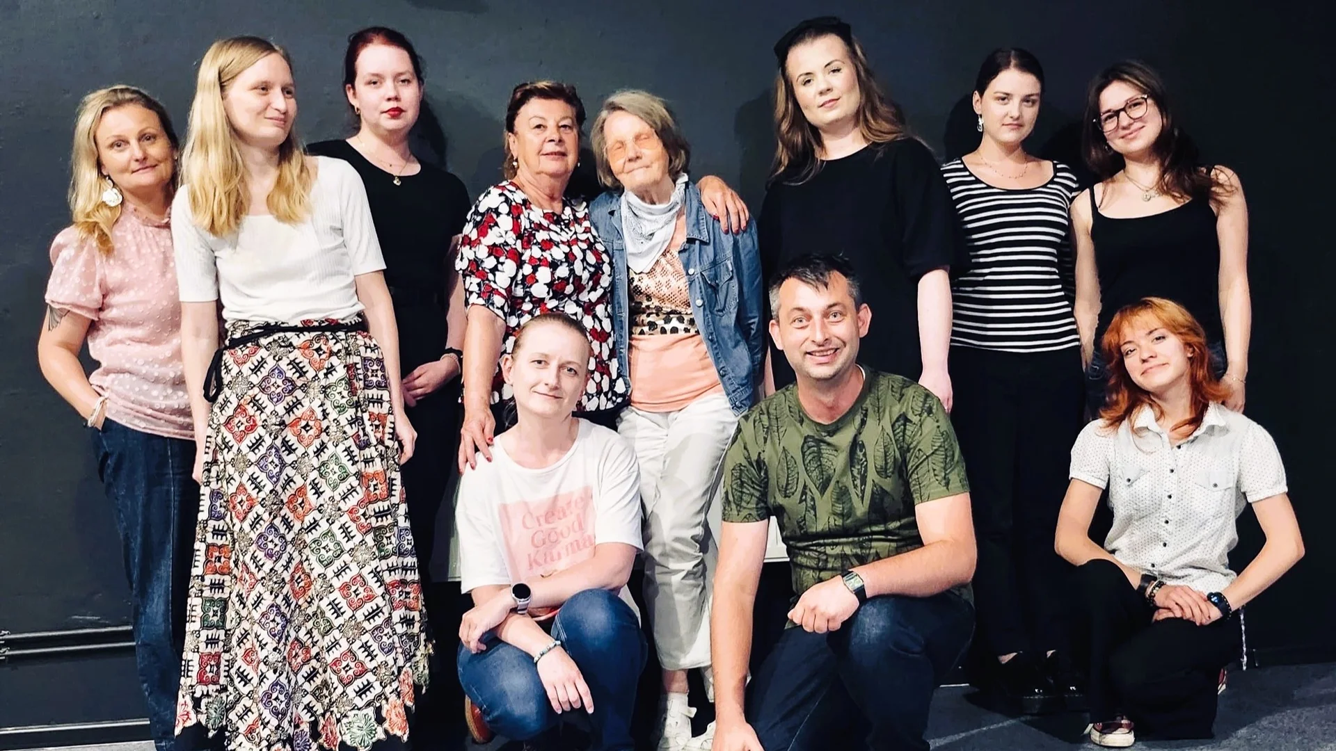 zdjęcie grupowe wolontariatu Teatru Współczesnego w Szczecinie. Z przodu kucają trzy osoby, reszta stoi z tyłu. W środku dwie kobiety w wieku seniorskim, obejmują się. Zdjęcie wykonane w sali prób. Czarne ściany, szara podłoga.