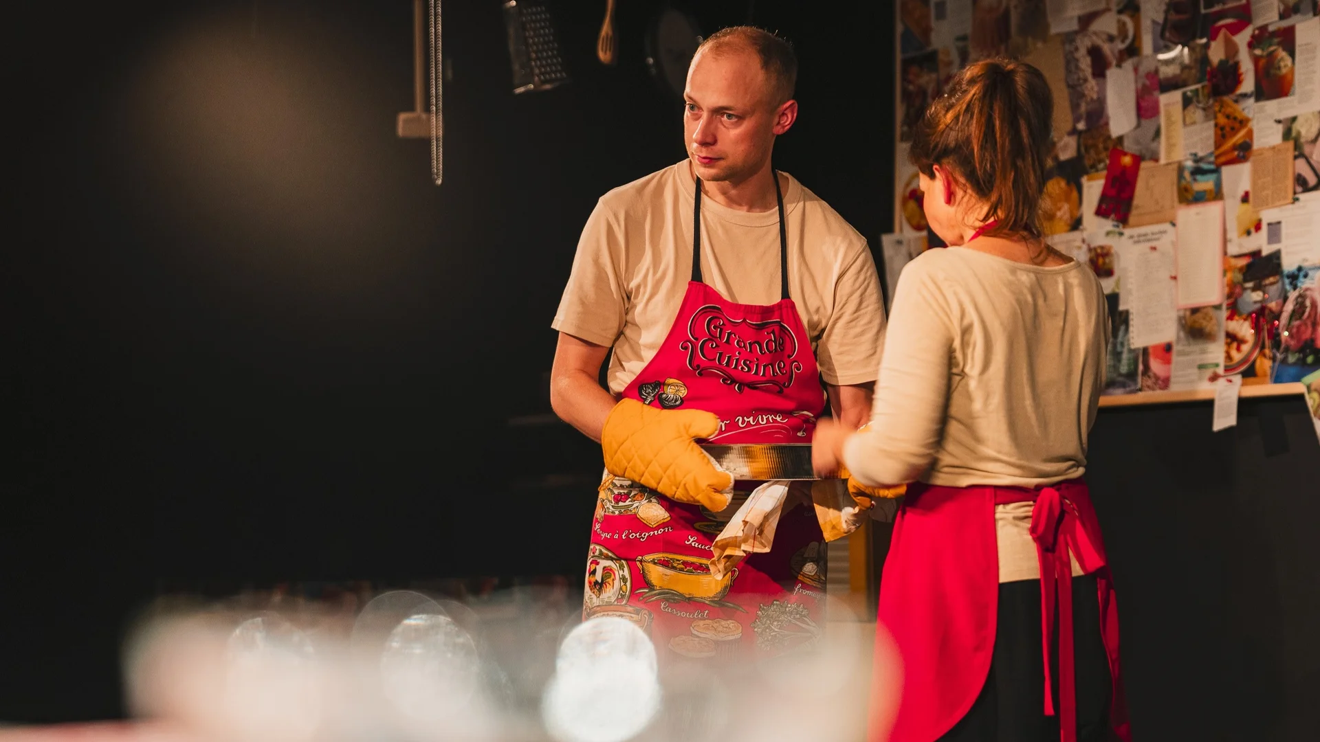 Aktorzy w kuchennych fartuszkach i rękawicach. Za nimi tablice korkowe z zawieszonymi przepisami wyciętymi z gazet. Na pierwszym planie coś niewyraźnego.