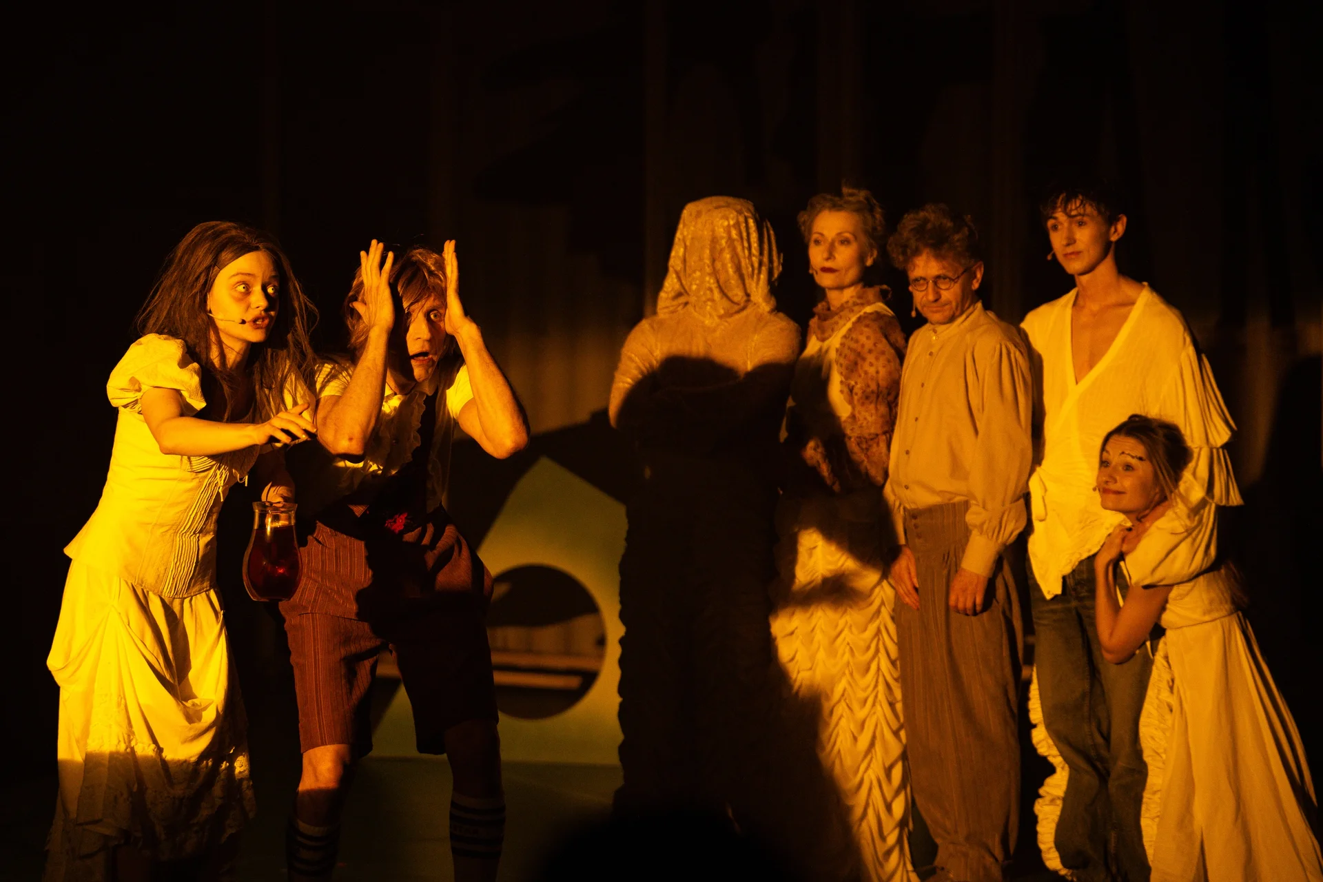 Grupa aktorów podświetlona żółtym światłem. Zdjęcia ma niepokojący charakter. Większość osób ma na sobie jasne ubrania. Na pierwszym planie mężczyzna, który trzyma się dłońmi za skronie.