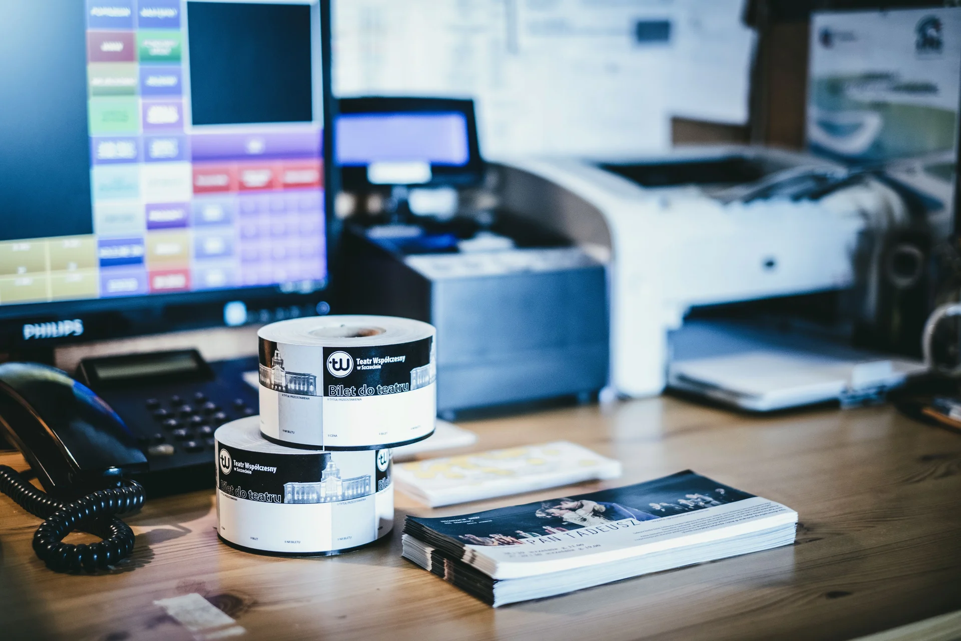 Zdjęcia kasy biletowej teatru. Na fotografii widoczne biurko, a na nim fragment ekranu komputera, sprzęt biurowy - telefon, drukarka i kasa. Na środku blatu  rolki i blankiety biletów do teatru.