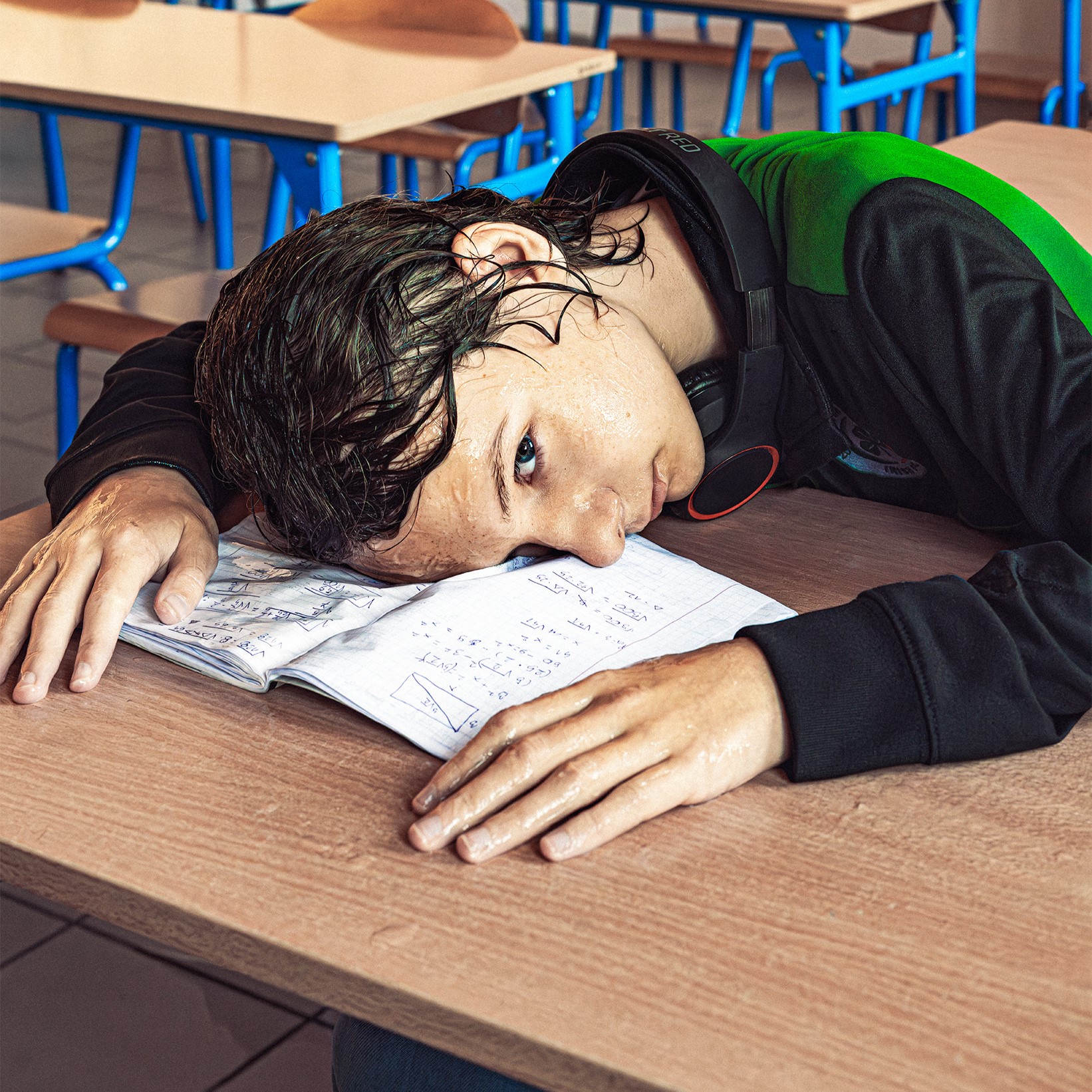 Młody chłopak z mokrymi włosami i w mokrych ubraniach trzyma głowę i dłonie na ławce. Pod buzią ma mokry zeszyt do matematyki. Zdjęcie zrobione jest w sali szkolonej, za chłopcem widać fragmenty ławek. 