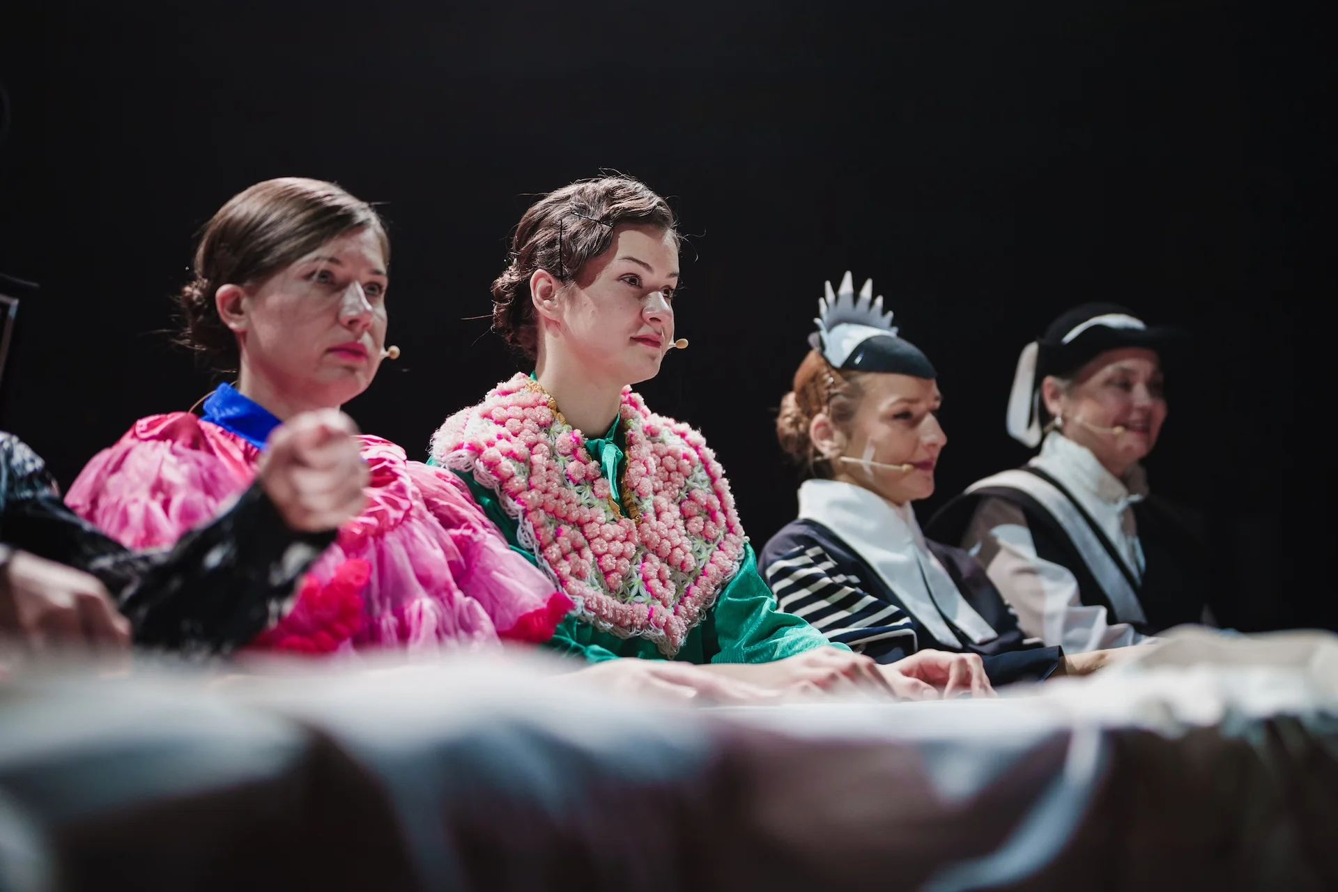 Cztery kobiety siedzące przy stole. Dwie z nich mają na sobie bardzo kolorowe kostiumy, a dwie kolejne czarno-białe i nakrycia głowy.