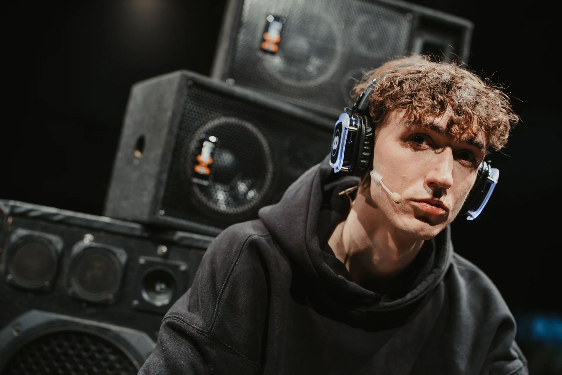 Chłopak z kręconymi włosami, w słuchawkach i czarnej bluzie w kapturem na tle głośników.