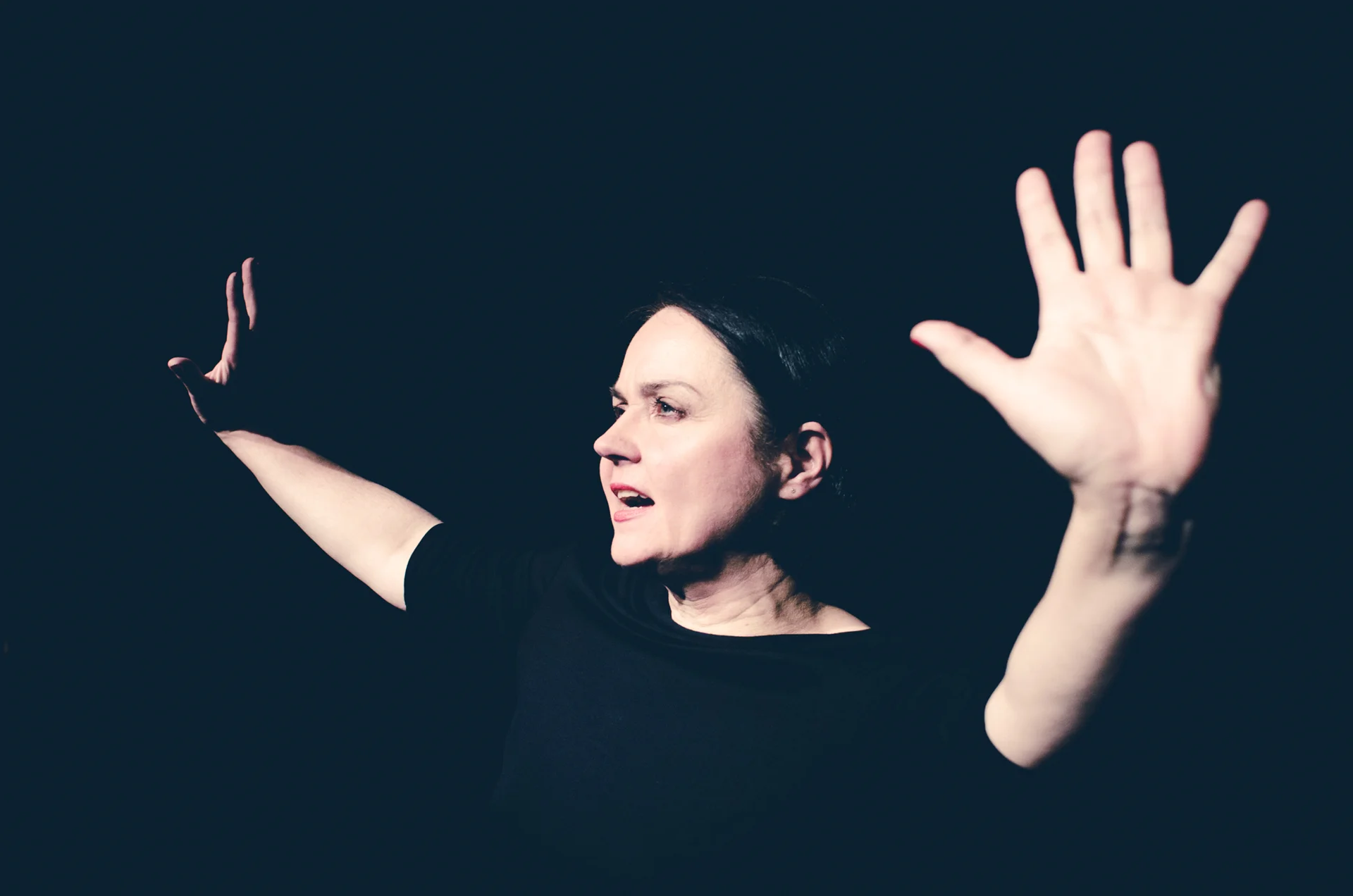 Kobieta w czarnej bluzce na czarnym tle. Oznacza się tylko jej twarz i dłonie, wyciągnięte do góry.