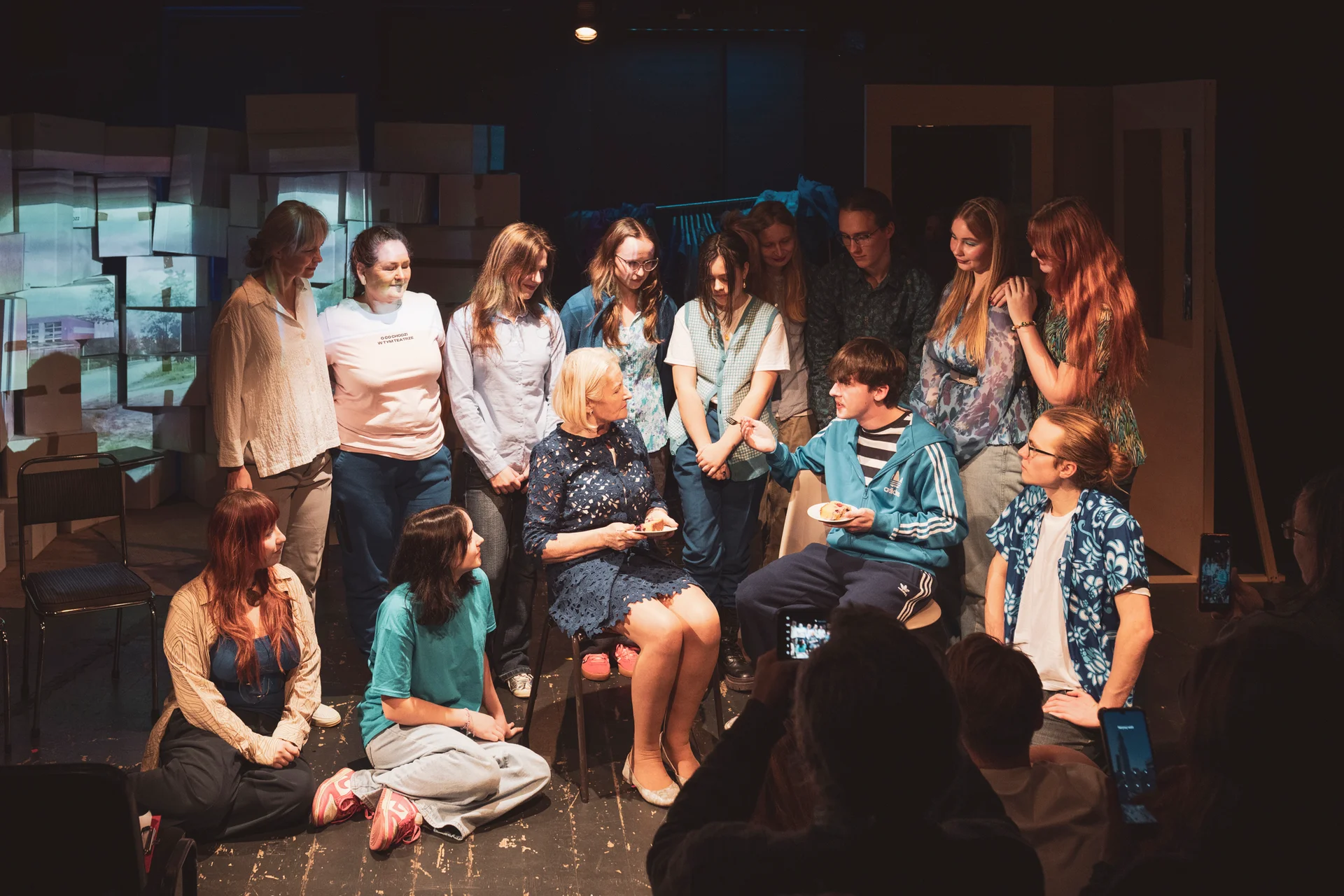 Międzypokoleniowa grupa osób na scenie. W środku młody mężczyzna i kobieta w starszym wieku siedzą na krzesłach. Kilka osób siedzi po bokach. Reszta stoi w tle.