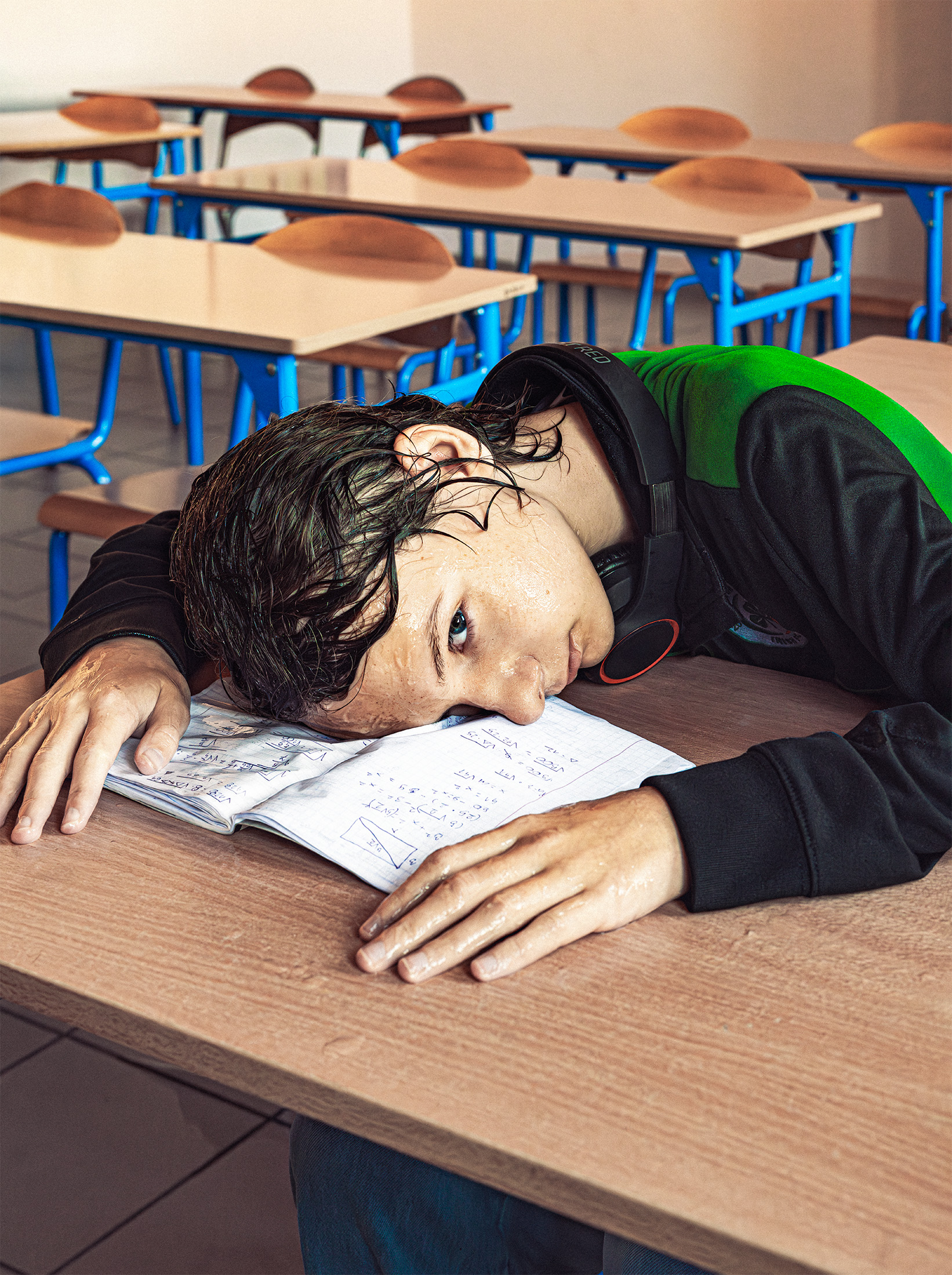 Młody chłopak z mokrymi włosami i w mokrych ubraniach trzyma głowę i dłonie na ławce. Pod buzią ma mokry zeszyt do matematyki. Zdjęcie zrobione jest w sali szkolonej, za chłopcem widać fragmenty ławek. 