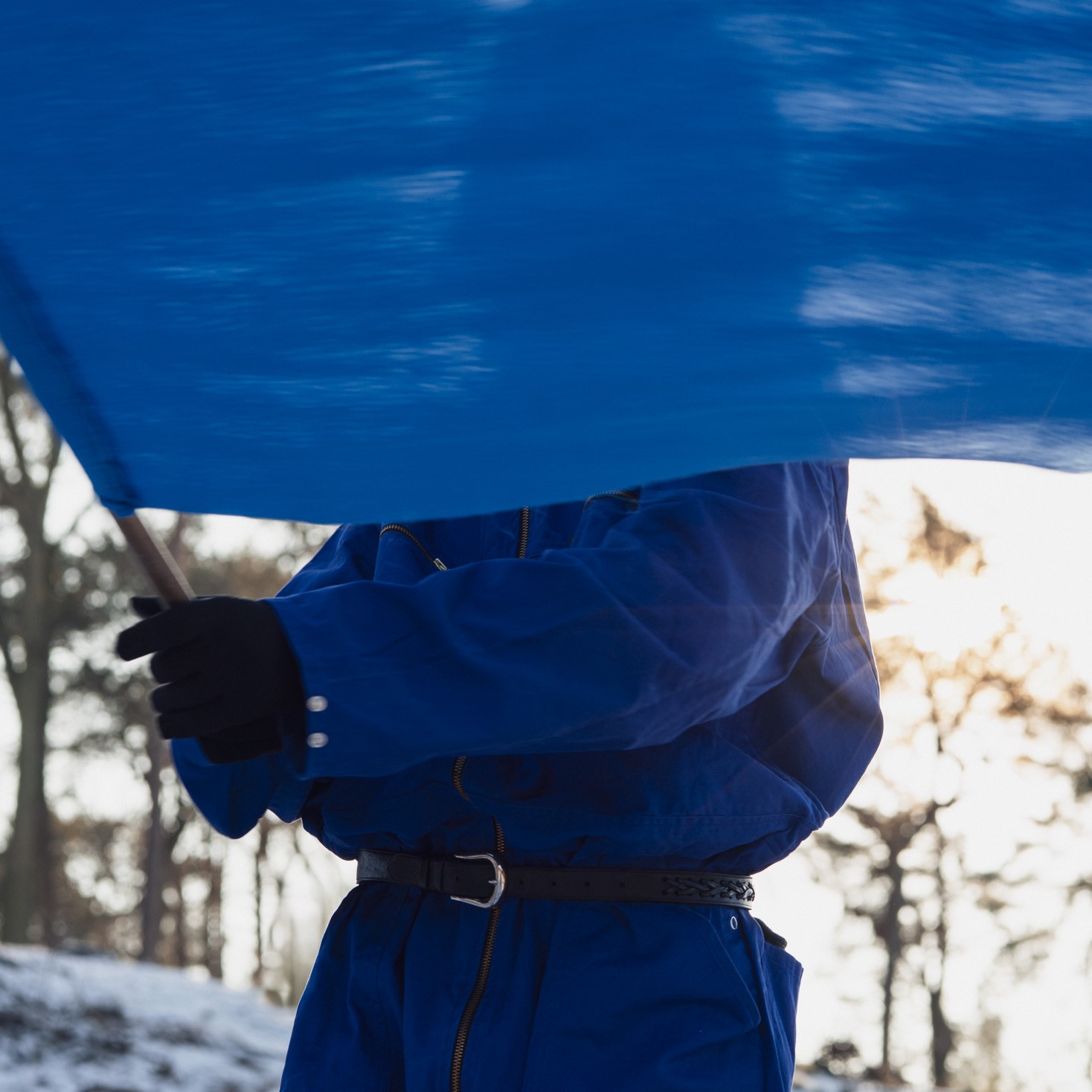 chłopak w niebieskim kombinezonie. Ma on przed sobą niebieską flagę, która zasłania jego twarz. W tle zimowy krajobraz - drzewa bez liści, fragment wzgórza ze śniegiem.