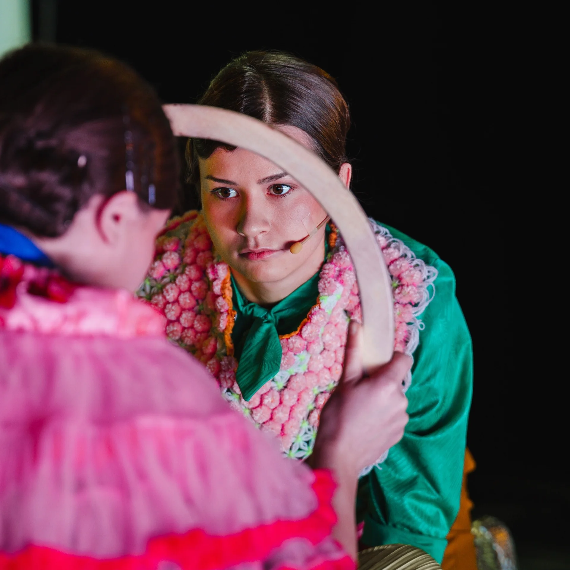 Twarz młodej kobiety, przed nią obręcz, trzymana przez drugą kobietę, którą widzimy tyłem. Obie mają ciemne włosy i mocno kolorowe bluzki na sobie.