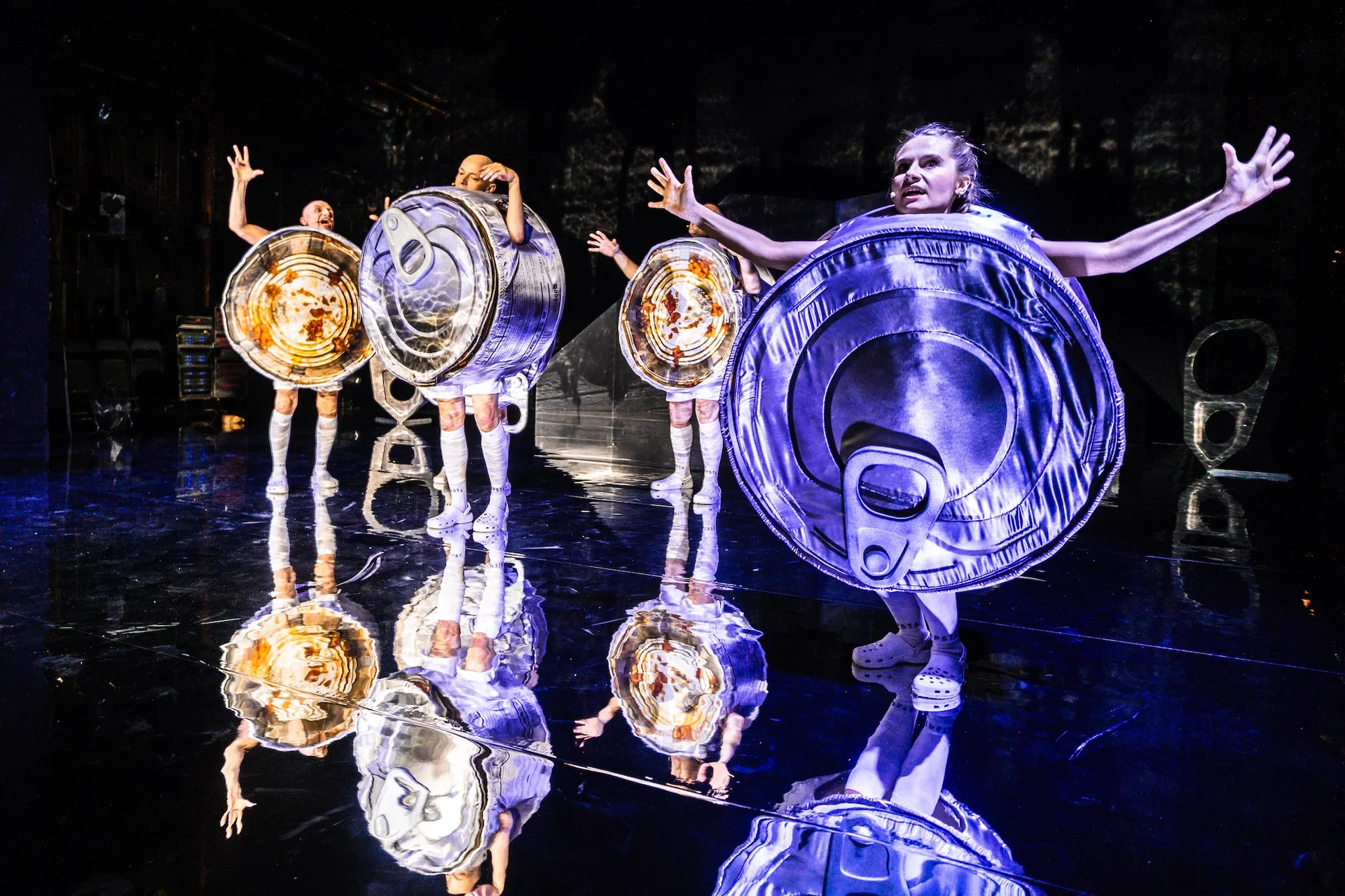 Na pierwszym planie kobieta w stroju puszki paprykarza szczecińskiego. Ma wyciągnięte w boki dłonie. Za nią kilka osób, również w strojach aluminiowych puszek. Podłoga jest lustrzana, widzimy w niej ich odbicie.