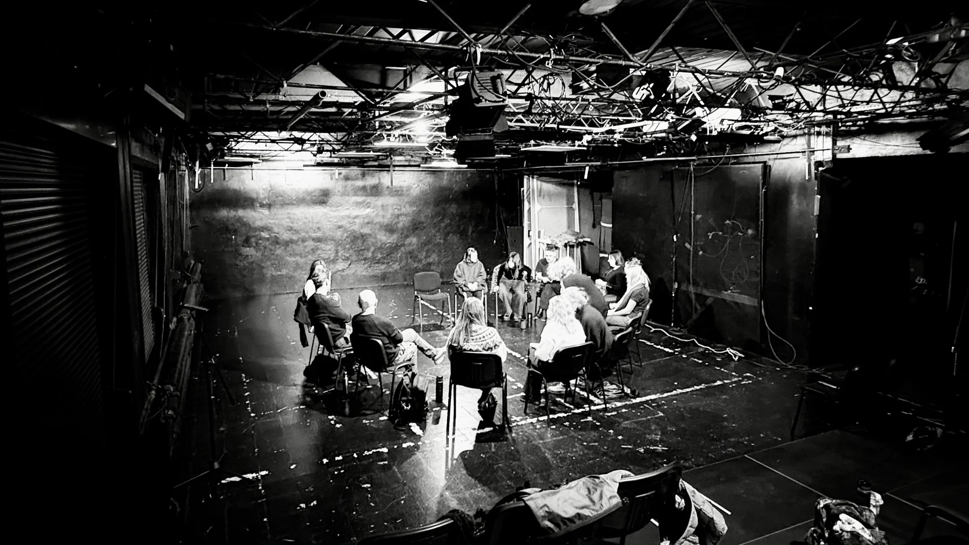 czarno-białe zdjęcie ze Sceny Nowe Sytuacje. Grupa aktorów i realizatorów siedzi na krzesłach w kole. 