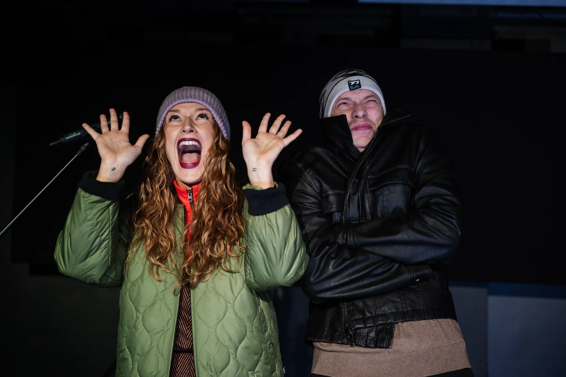 Zdjęcie ze spektaklu Nie twoja kolej, kochanie. Kobieta i mężczyzna w kurtkach zimowych i czapkach. Mężczyzna wygląda jakby  było mu zimno, ma założone ręce na siebie. Kobieta unosi dłonie ku górze, wygląda jakby coś wykrzykiwała, ma szeroko otwarte usta.