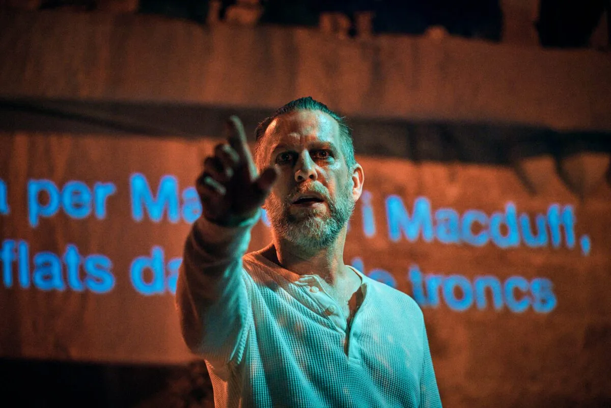 Mężczyzna w jasnej koszuli skazuje palcem przed siebie. Za nim na niebiesko wyświetlane są napisy.