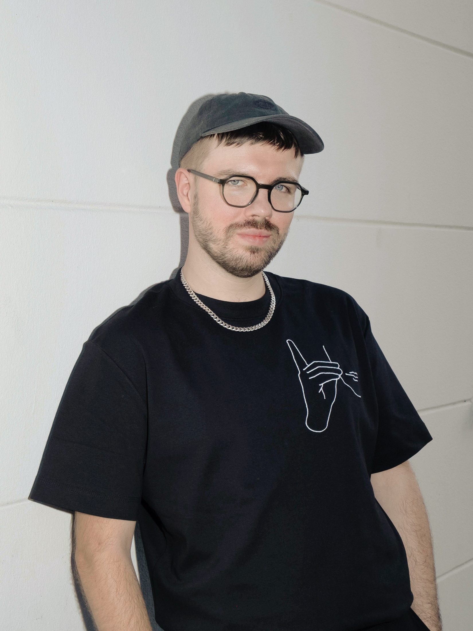 Mężczyzna stojący na tle białej ściany. Na głowie ma szarą czapkę z daszkiem i czarne okulary. Ubrany w czarną koszulkę z krótkim rękawem z wyhaftowaną białą dłonią.