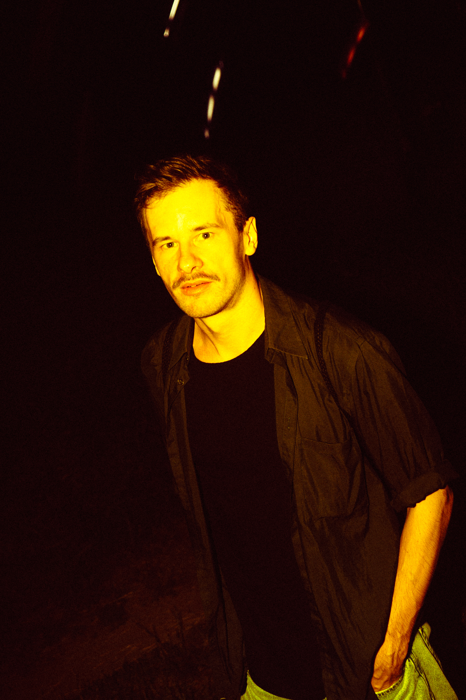 Portret mężczyzny ubranego w rozpiętą czarną koszulę z podwiniętymi rękawami, pod spodem ma czarną koszulkę. Lewą rękę ma włożoną w kieszeń jasnych dżinsów. 