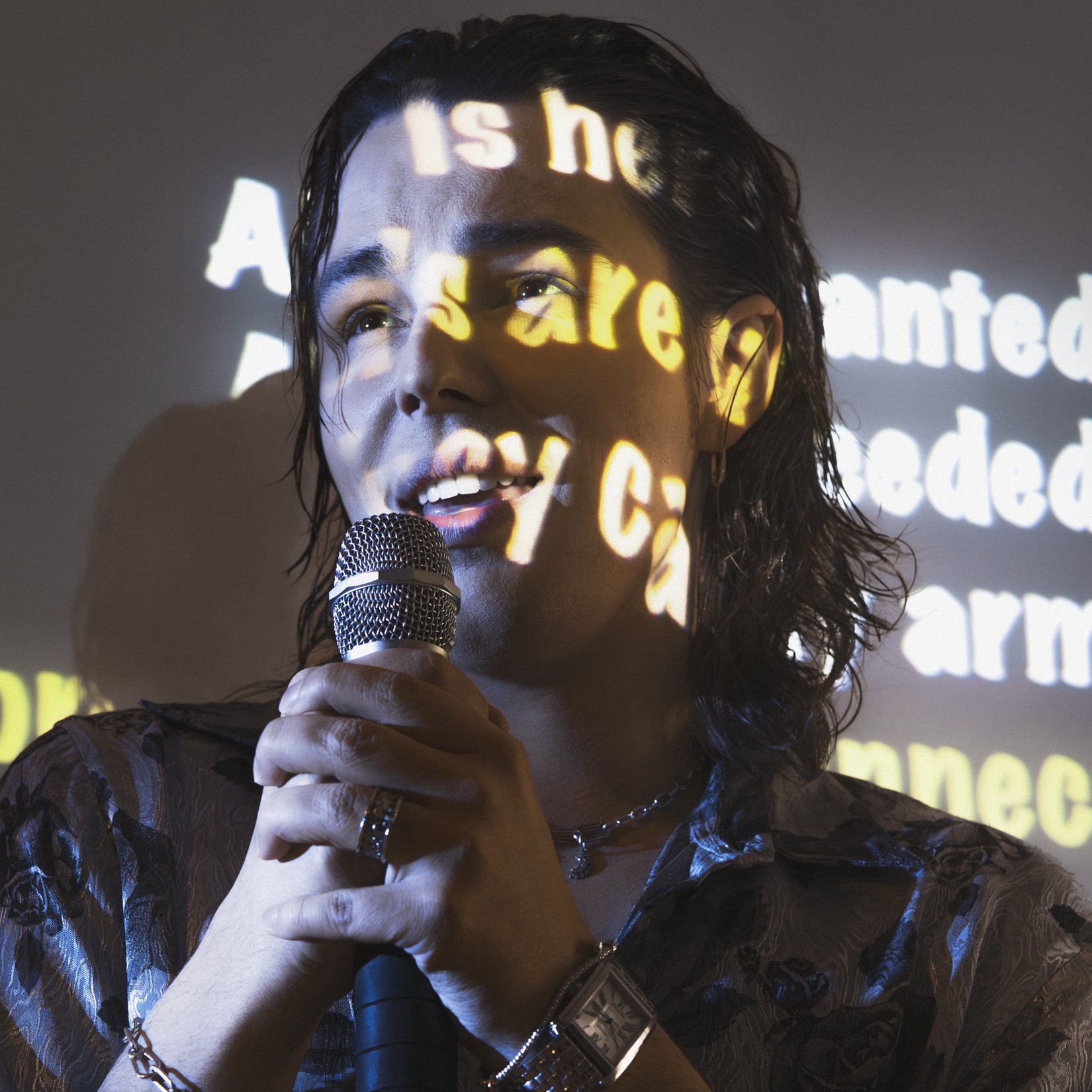 Młody mężczyzna śpiewa do mikrofonu. Na jego twarzy wyświetlane są słowa piosenki. 