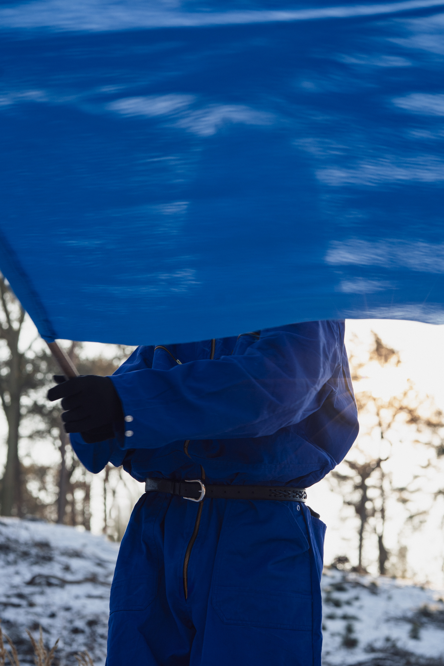 fot. Mila Łapko Osoba w niebieskim kombinezonie. Ma ona przed sobą niebieską flagę, która zasłania jej twarz. W tle zimowy krajobraz - drzewa bez liści, fragment wzgórza ze śniegiem.