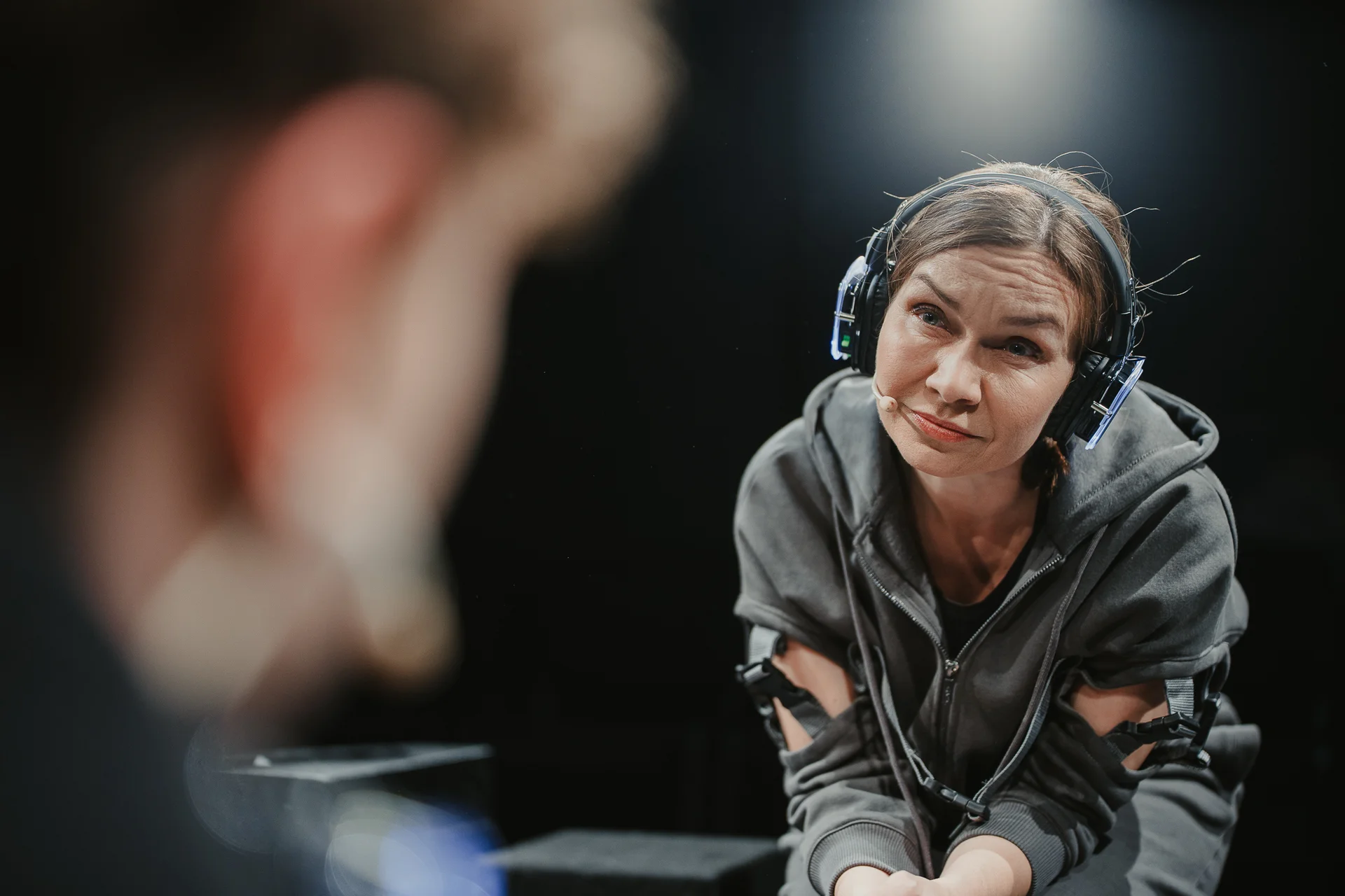 Kobieta w dużych słuchawkach nausznych. Ubrana w szarą bluzę z kapturem z wycięciami na ramionach. Pochyla się z miną sugerującą współczucie. Jej wzrok jest zwrócony w stronę rozmazanej postaci zajmującej pierwszy plan fotografii. 