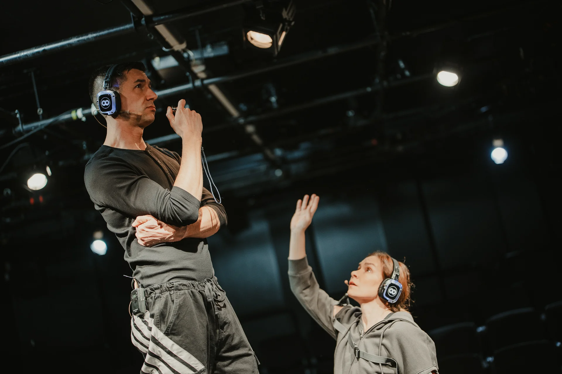 Mężczyzna w ciemnym sportowym ubraniu stoi z założonymi rękoma. Na głowie ma duże słuchawki nauszne. Jest zwrócony w prawą stronę. Przed nim klęczy kobieta z uniesioną w górę prawą ręką. Ubrana jest w szarą bluzę z kapturem, a na głowie ma duże słuchawki nauszne. 