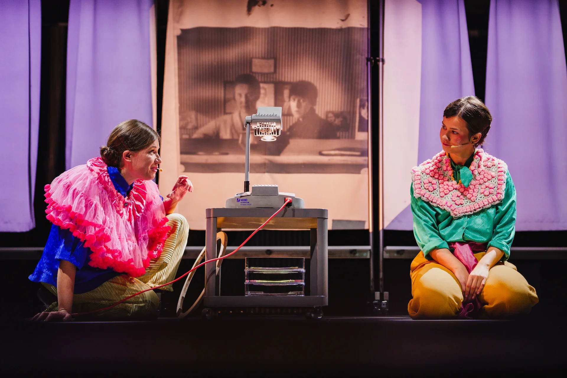 Dwie kobiety w kolorowych strojach siedzą na podłodze. Aktorka z lewej strony ubrana jest w granatową koszulę, żółte spodnie, a na szyi ma wielki, falbaniasty różowy kołnierz. Zwrócona jest ku drugiej kobiecie. Kobieta po prawej stronie zdjęcia patrzy w skupieniu na swoją towarzyszkę. Ubrana w żółte spodnie, turkusową koszulę i biało-różowy kołnierz zrobiony na szydełku. 