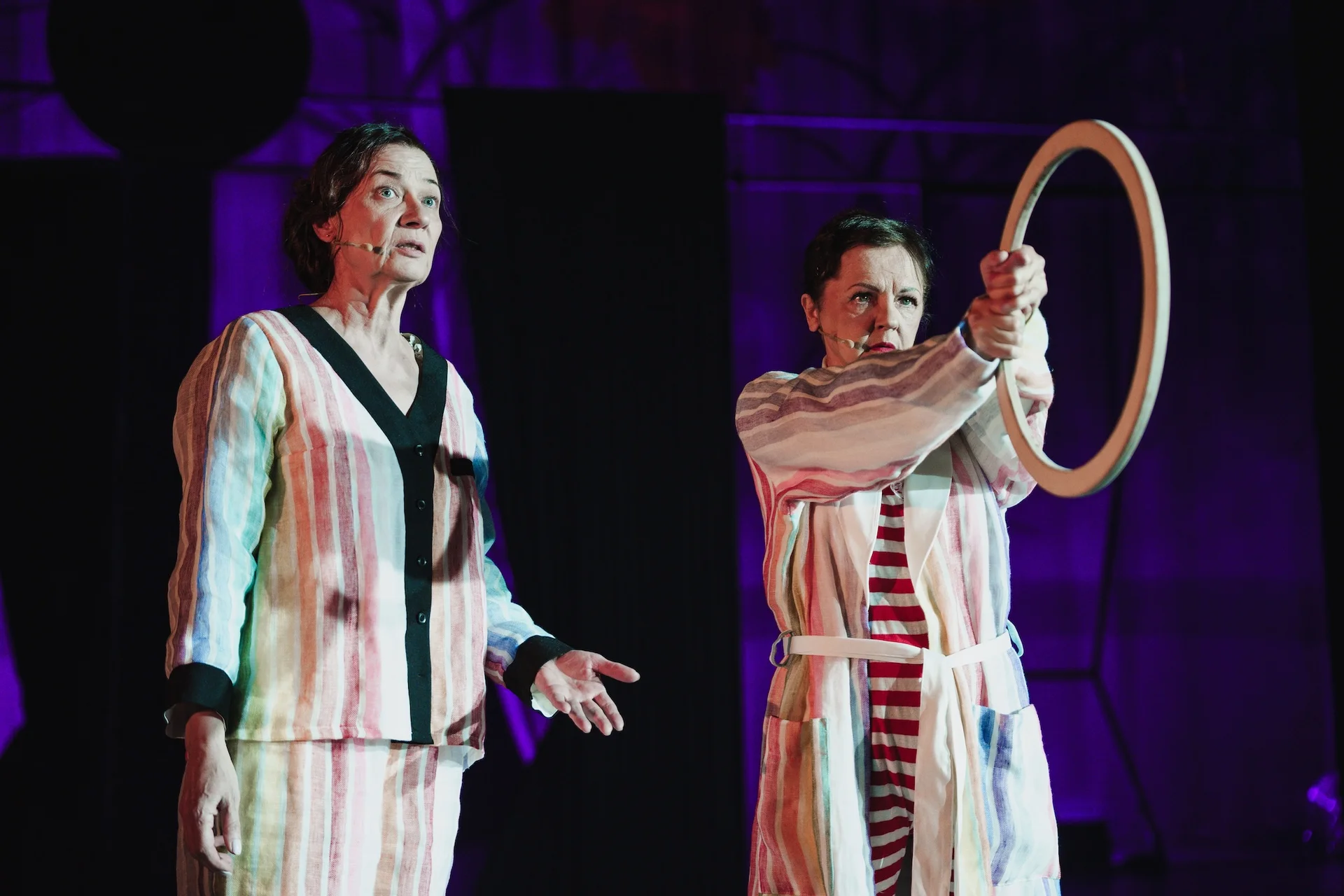 Dwie kobiety stoją z dwóch stron zdjęcia. Aktorka po lewej stronie ubrana jest w pastelową pasiastą piżamę z czarnymi lamówkami. Kobieta po prawej ubrana jest w pasiasty szlafrok zakrywający biało-czerwony strój w paski. Ręce ma wyciągnięte przed siebie, a w nich trzyma drewnianą obręcz. 