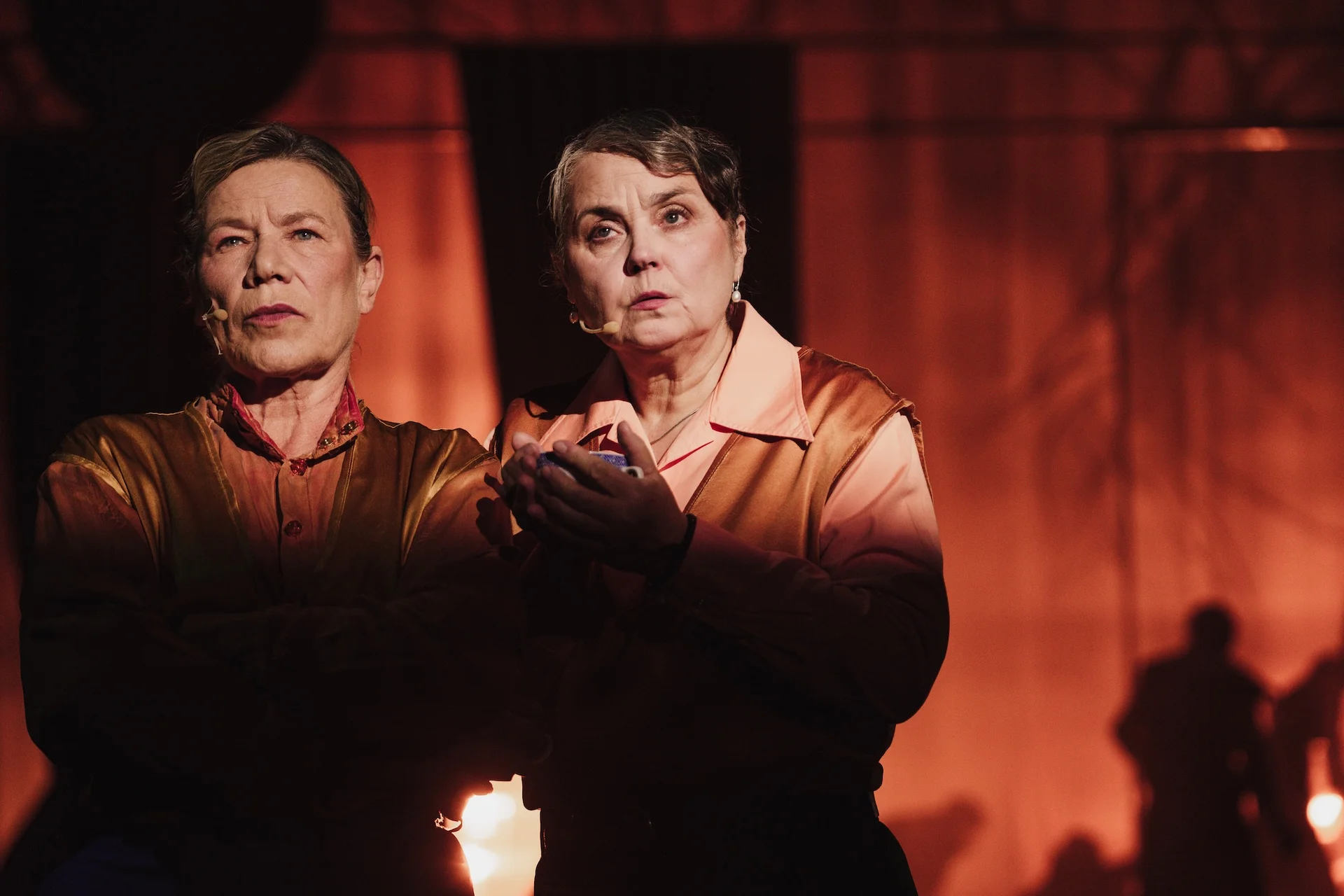 Dwie kobiety stoją ubrane w odcienie złota i różu. Patrzą w dal z poważnymi minami. 