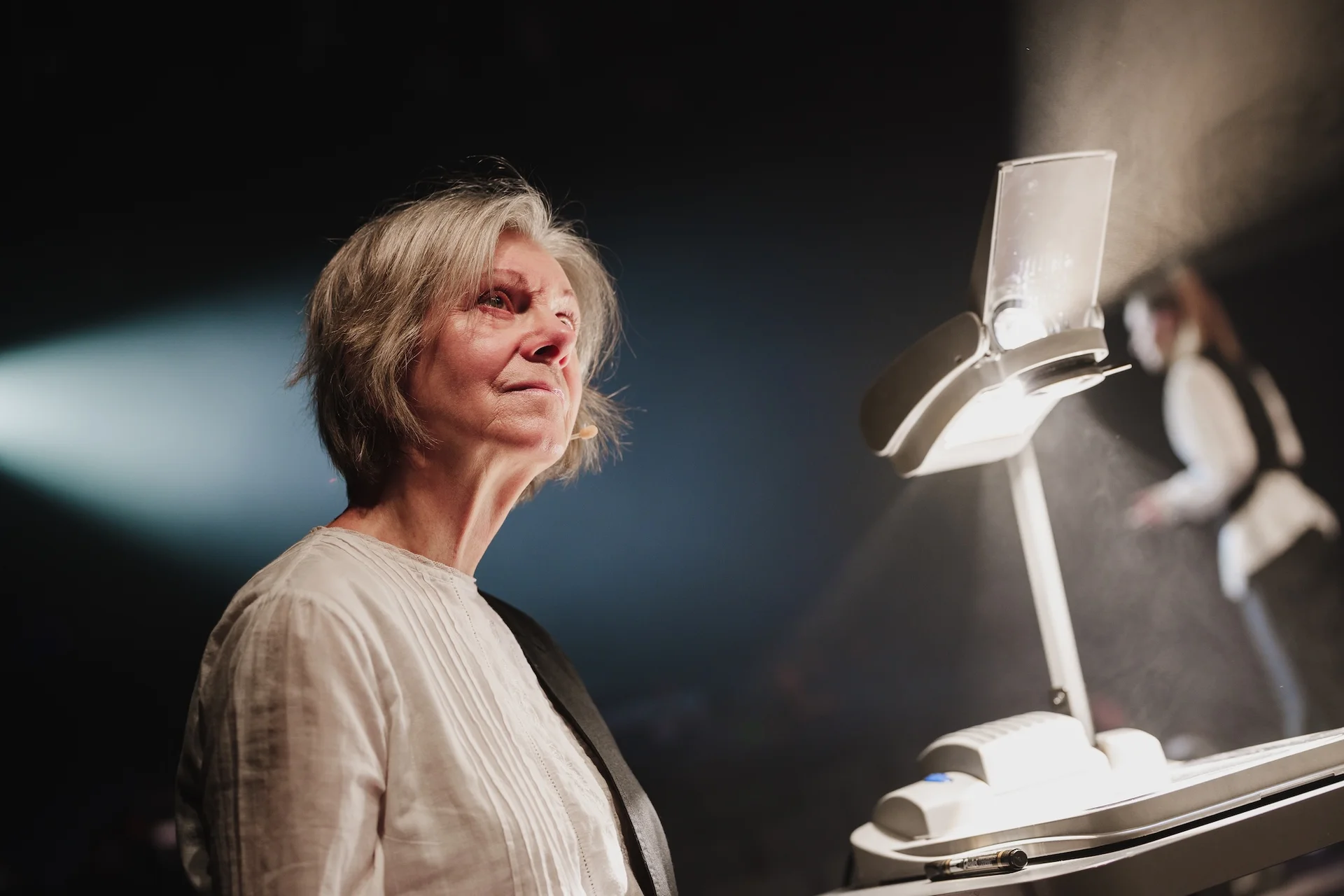 Kobieta ubrana w białą koszulę spogląda z nostalgią na obraz z projektora, który wyświetlany jest poza zdjęciem. 