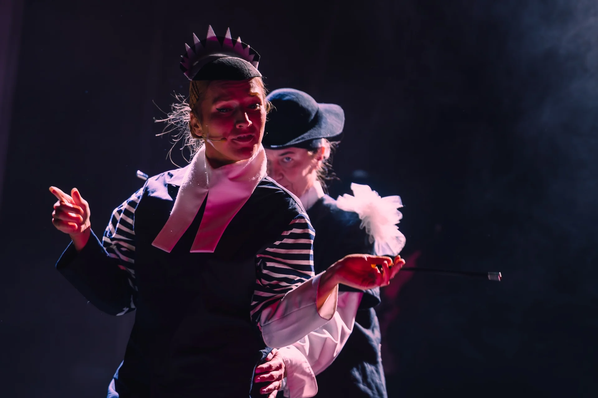 Kobieta w czarno-białym stroju stoi z cygaretką w lewej dłoni. Za nią znajduje się druga aktorka, z lewą dłonią położoną na jej biodrach. 