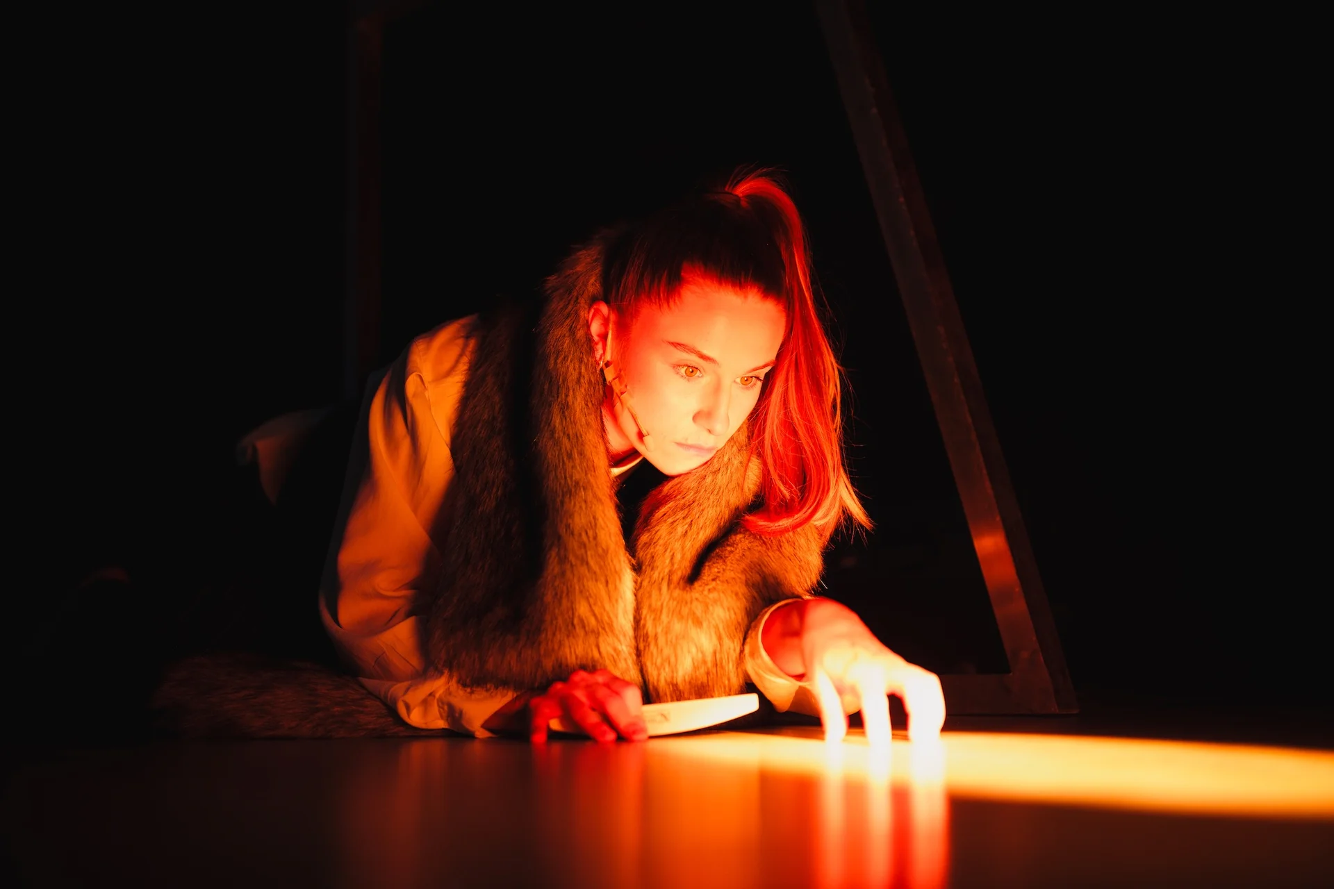 Kobieta leżąca na podłodze podpiera się na łokciach. Wzrok ma skupiony na smudze światła przed nią. Na sobie ma ubrane futro, włosy ma spięte w wysoką kitkę. 