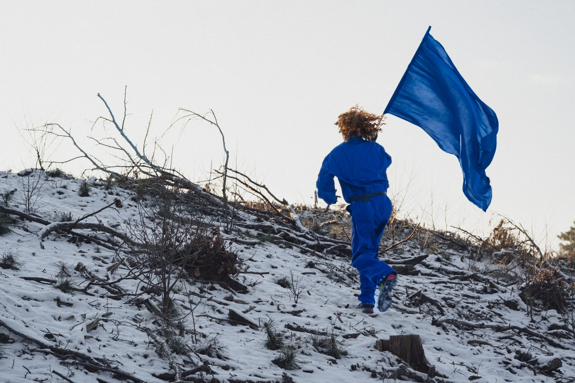 Młody chłopak w niebieskim uniformie z niebieską flagą wbiega pod górę. Ma rude kręcone włosy. Wokół leży delikatny śnieg.