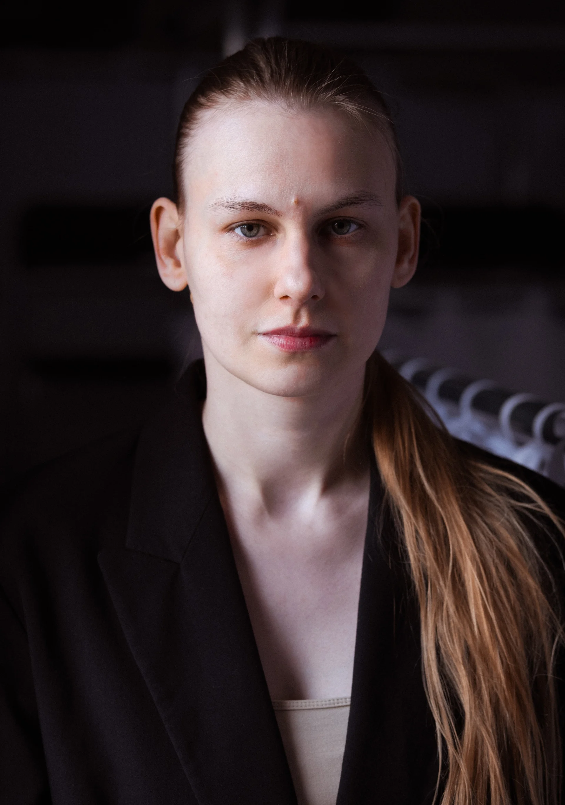 Portret kobiety patrzącej wprost. Ma długie włosy spięte w kitkę opadającą na lewe ramię. Ubrana jest w czarną marynarkę i jasno-beżową podkoszulkę. 