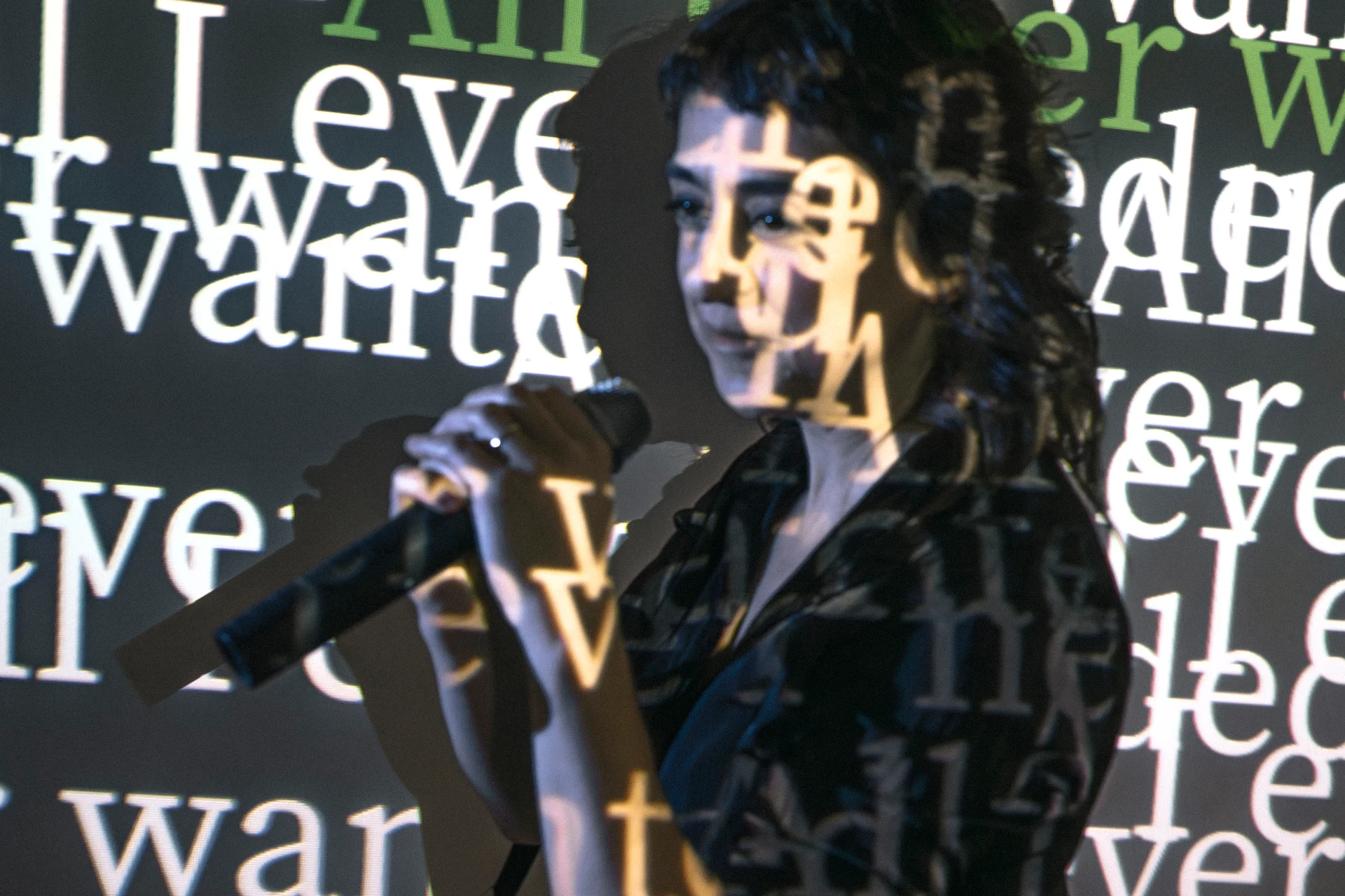 fot. Mila Łapko Zdjęcie kobiety trzymającej mikrofon, ubranej w ciemną koszulę. Pokazana jest na tle ciemnego ekranu, na którym wyświetlane są nachodzące na siebie słowa. Napisy wyświetlane są też na kobiecie.